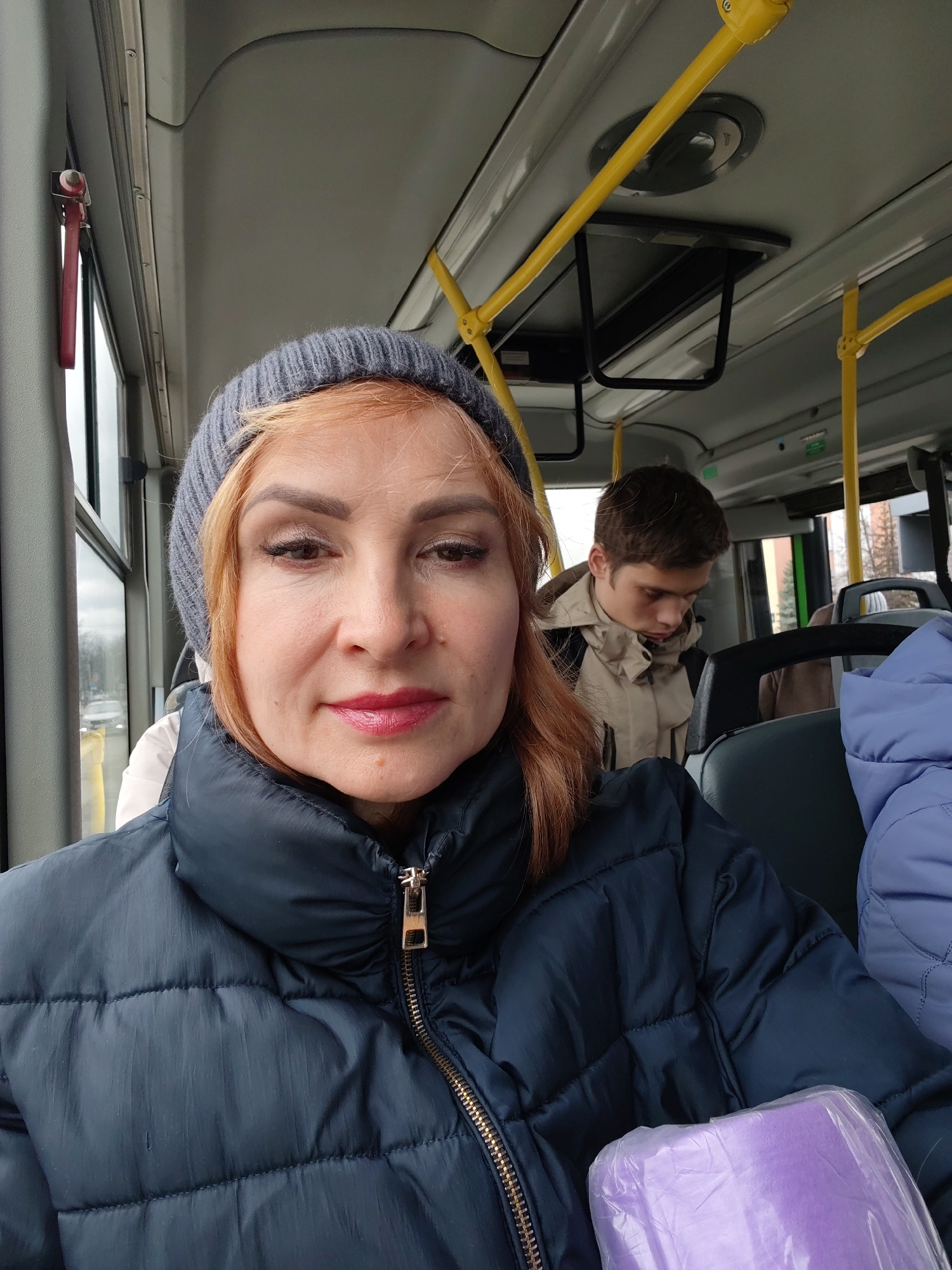 A woman with reddish-blonde hair and a gray knit hat is sitting on a bus. She is wearing a navy blue puffer jacket. In the background, a young man with short brown hair is sitting in a beige jacket. There are yellow handrails visible inside the bus. The location is indicated as Chaikovsky, Russia.