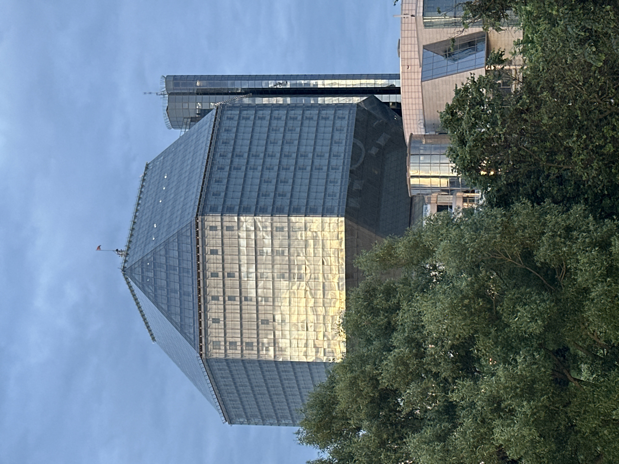 The image displays the National Library of Belarus in Minsk, Belarus. The primary subject is a large, multi-faceted building with a distinctive geometric shape, predominantly clad in glass. The left and front-facing facets of the building exhibit a strong golden-yellow reflection, indicative of direct sunlight, contrasting sharply with the cooler blue-grey tones on the right-facing facets. The upper structure of the main building tapers into a glass pyramid, from the apex of which a small red flag is visible. The facade is segmented into numerous square or rectangular panels. To the right of the main library, a taller, slender skyscraper, also clad in glass, is partially visible against the sky. In the foreground, dense green foliage from trees partially obscures the lower sections of the main library building and other modern, lower-rise structures with glass and light-colored facades. The sky above is a uniform blue-grey, indicating an overcast condition or a specific time of day. No individuals or specific actions are discernible within the frame.