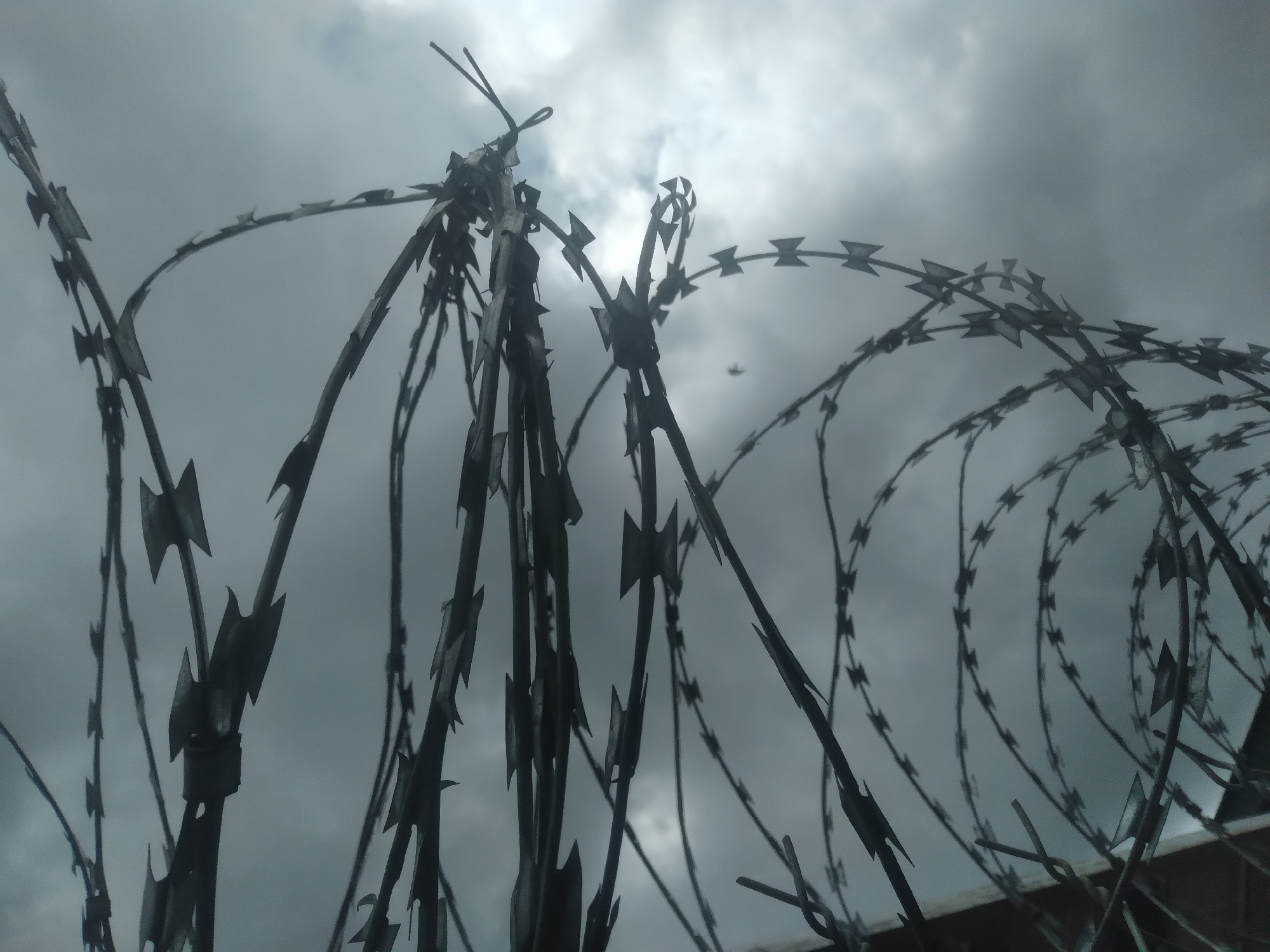 This image displays a close-up view of barbed wire, likely a security fence, against a cloudy, overcast sky. The barbed wire is arranged in coiled and linear patterns, with sharp, pointed barbs clearly visible. The sky is a uniform gray, suggesting either a heavily clouded day or dawn/dusk. There is a hint of a dark structure or wall in the lower left portion of the image, which the barbed wire appears to be attached to. No people are visible in the frame. The overall impression is one of security or containment. The specific location in the city of Durimi, Nigeria, cannot be determined from the visual information provided in the image alone.