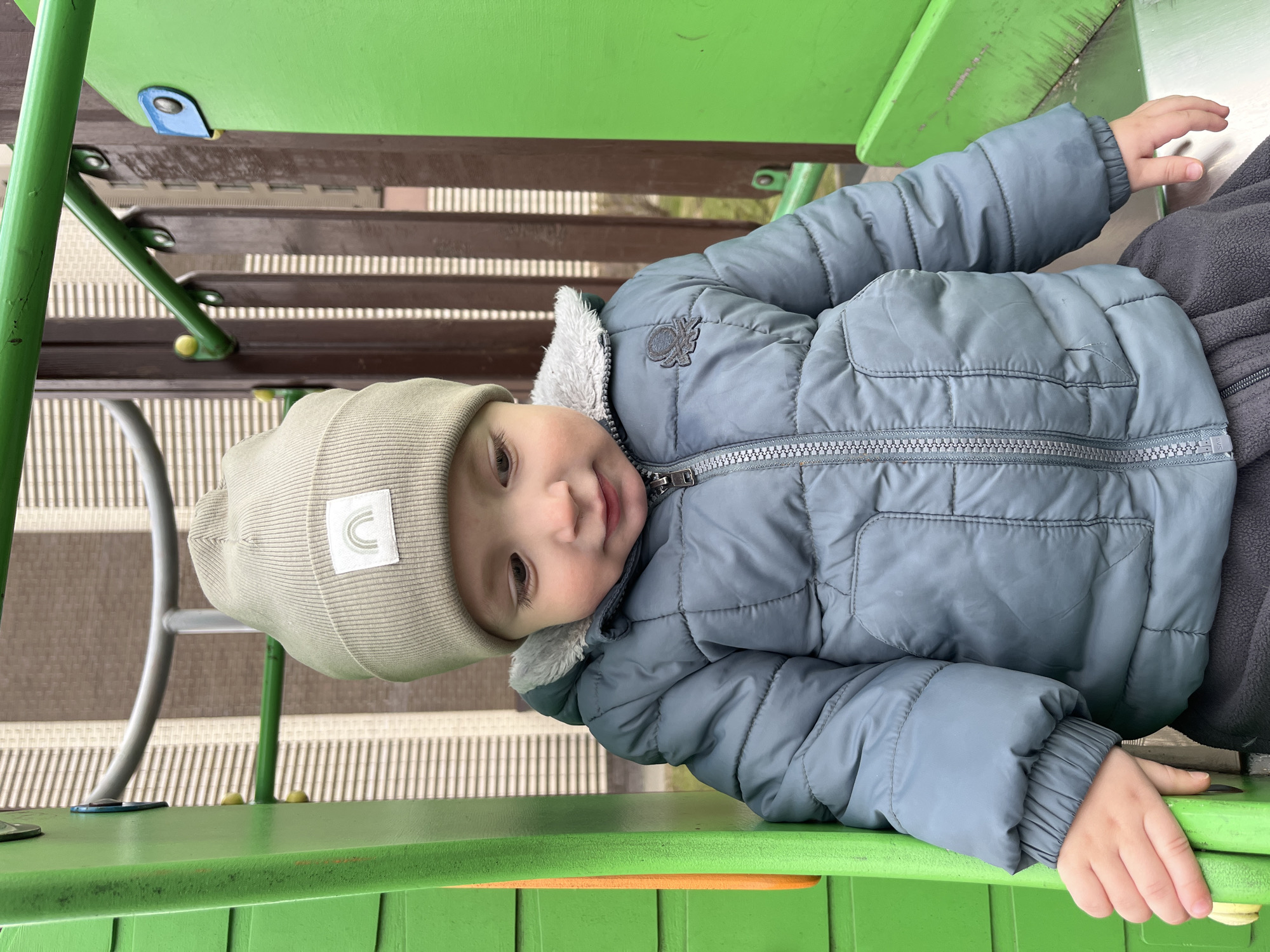 A young child, likely a toddler, is captured sitting on a bright green surface of a playground structure. The child looks directly towards the camera with a gentle, curious expression and a slight smile. They are dressed in warm clothing, including a light blue-grey puffy winter jacket featuring a light grey faux fur lining at the collar and a small, dark embroidered emblem on the left chest. The child also wears a light beige ribbed beanie hat adorned with a white rectangular patch that displays a light grey-green curved rainbow or arch symbol.
The setting is an outdoor playground, identified as being in Akulovo (Gp Kubinka), Russia. The green surface the child is on is part of a larger play structure, with brown slatted sections and green and silver metal supports visible in the background, indicative of a multi-level play area. The natural lighting suggests it is daytime. The child's hands are resting on or near the green surface. The image conveys a calm and cheerful atmosphere, focusing on the child's engaging gaze. Aside from the symbols on the clothing, no other visible text is present.