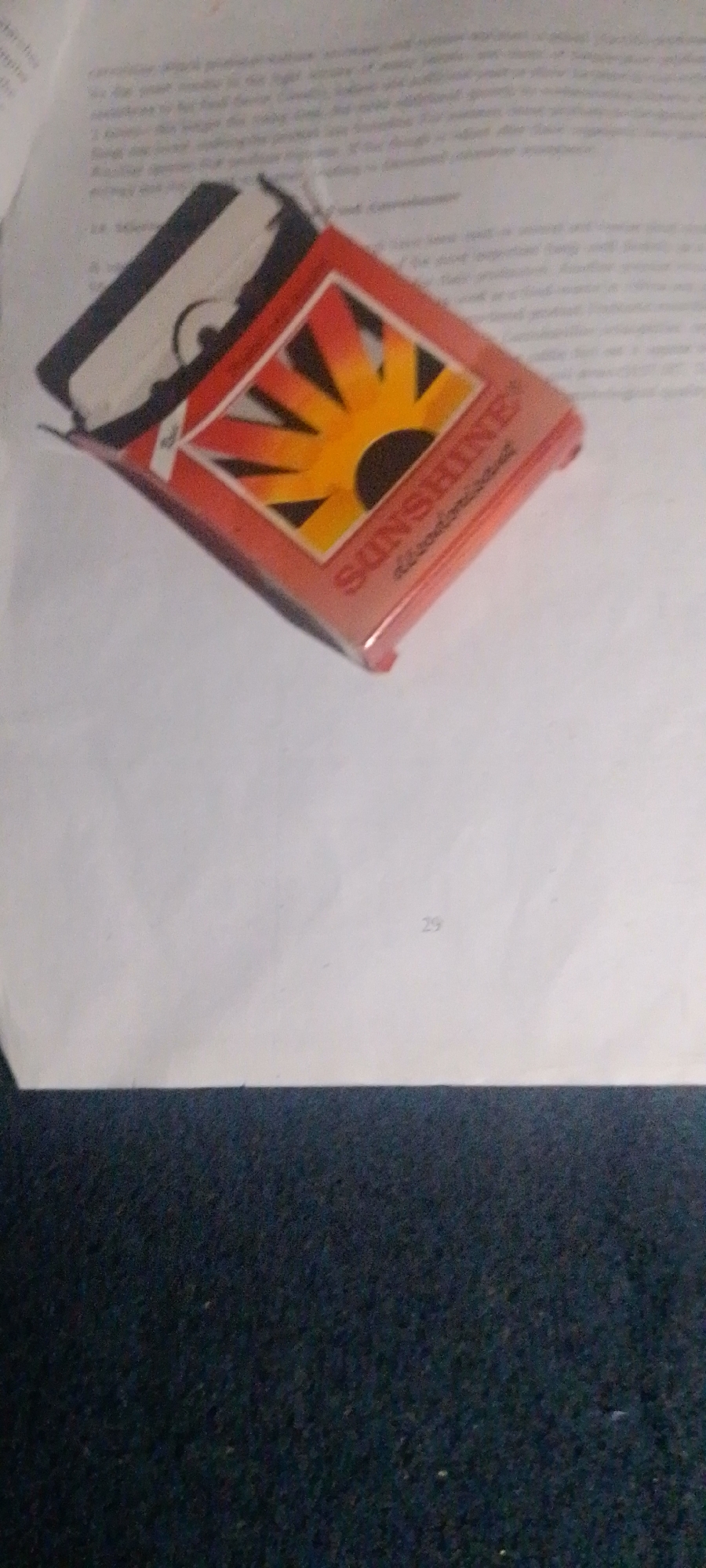 A close-up, slightly angled shot shows a partially opened red and white cigarette pack with a sunburst logo and the word "SUNSHINE" visible. The pack is lying on a white sheet of paper that has some text printed on it. Below the paper, a dark, textured surface, possibly carpet or fabric, is visible. The image does not contain enough information to determine its location within the city of Jalingo, Nigeria.