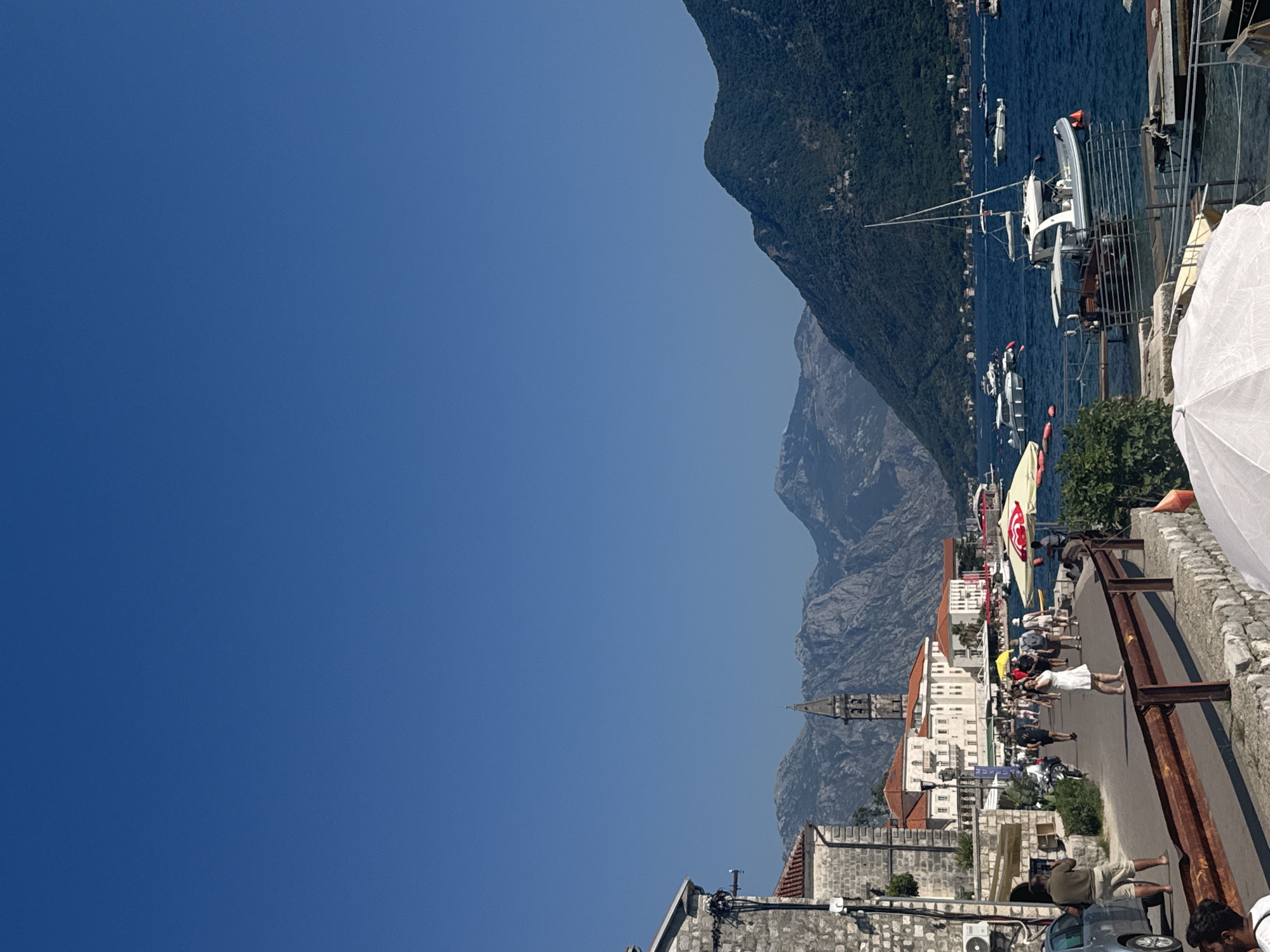 A bright, sunny day unfolds in Perast, Montenegro, with a clear blue sky dominating the upper half of the frame. The scene captures a waterfront promenade lined with stone buildings and a paved walkway. To the left, a stone building with red-tiled roofs is visible. A man stands on the walkway, holding a camera and seemingly filming. Further along, a group of people stroll, some in light clothing, suggesting a warm climate. A few motorcycles are parked near the buildings.
The promenade overlooks a bay filled with several boats, ranging from small motorboats to a larger white sailboat with a mast. The water is a deep blue, reflecting the sky. In the background, a picturesque mountain range rises, its slopes covered in green vegetation. The peaks are rugged and distinct, adding a dramatic backdrop to the coastal town. A church steeple with a clock tower is prominent among the buildings. The overall atmosphere is one of a relaxed, tourist-friendly environment.