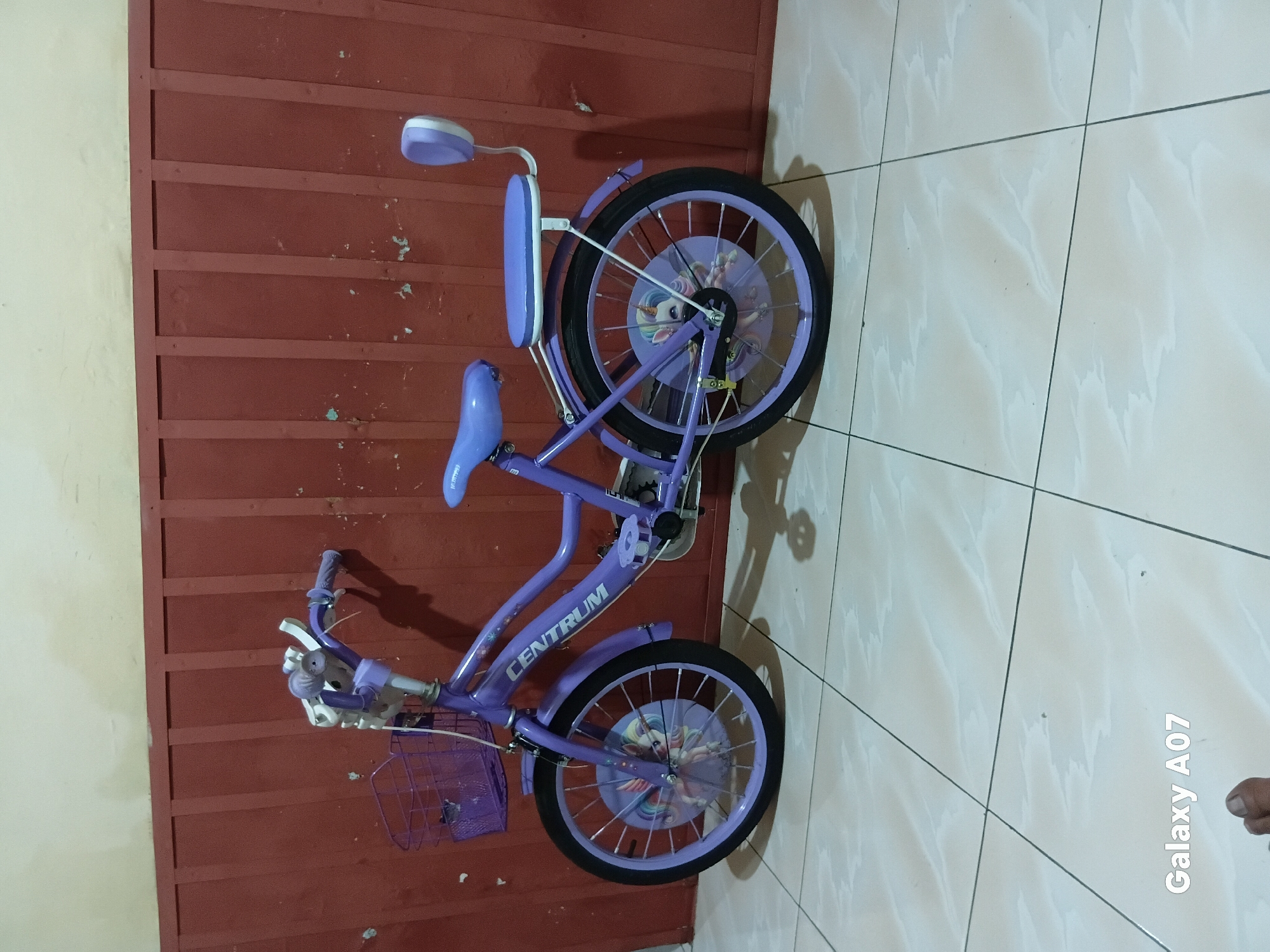 A purple child's bicycle with a front basket and a padded rear seat is parked on a tiled floor in front of a red wall. The bicycle has a "CENTRUM" logo on the frame and colorful unicorn decals on the wheel covers. The wall appears to be made of vertical wooden planks. The floor tiles are light-colored with a subtle pattern. The image has a watermark in the bottom left corner that reads "Galaxy A07". There is no visible indication that the image was taken in Batu, Indonesia, or any other specific city.