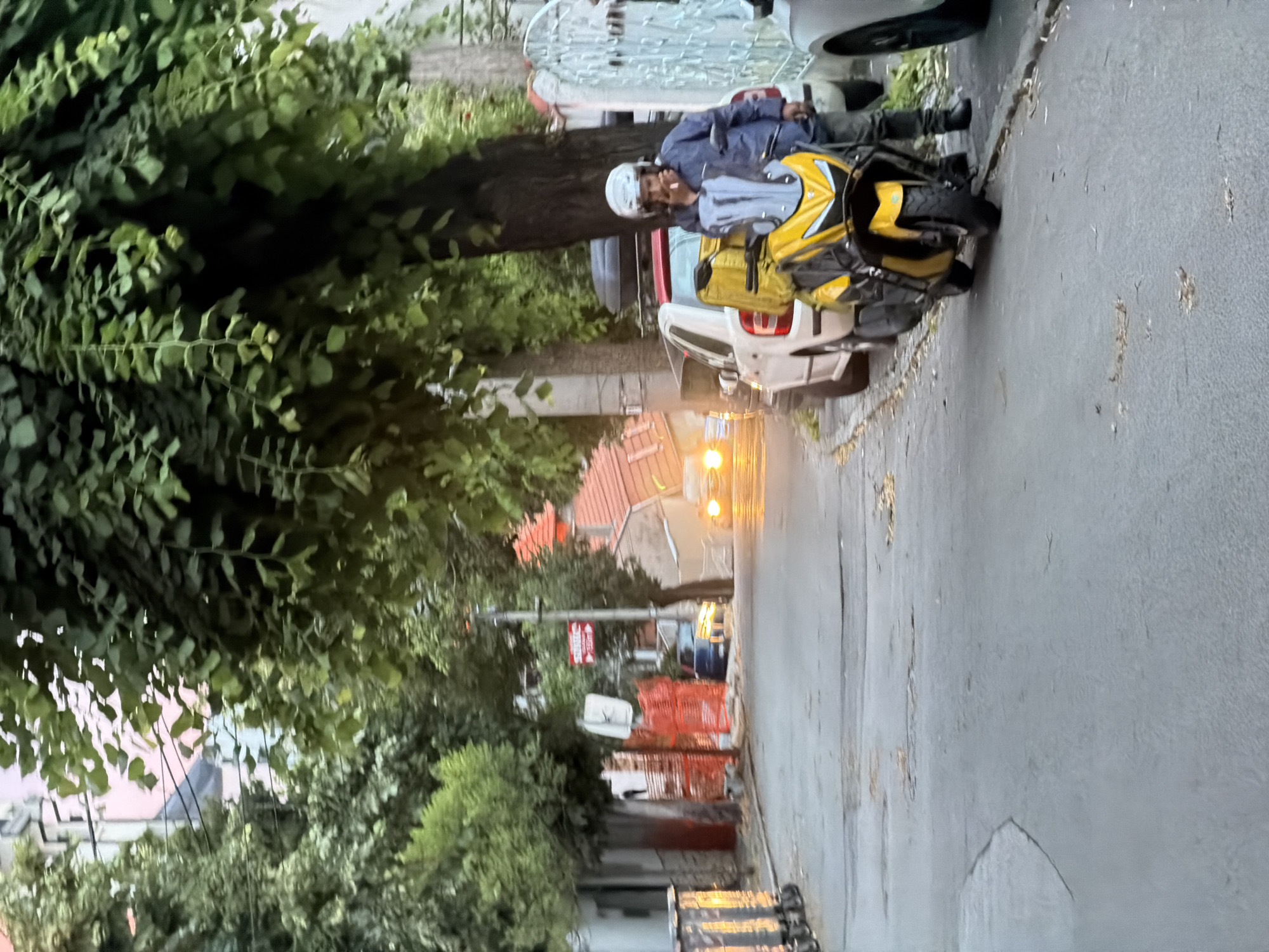 A person wearing a blue jacket, dark pants, black boots, and a white helmet is standing next to a yellow scooter on a paved street. The scooter has a large yellow storage box on the back and a gray windshield. The person is holding a cigarette in their right hand, and appears to be smoking. The street is lined with trees and buildings. There are cars parked along the side of the street, including a white car and a dark blue car. In the background, there are houses with orange roofs and construction fencing. The overall scene suggests an urban environment in Palilula Urban Municipality, Serbia, possibly during dusk or early morning given the lighting.
