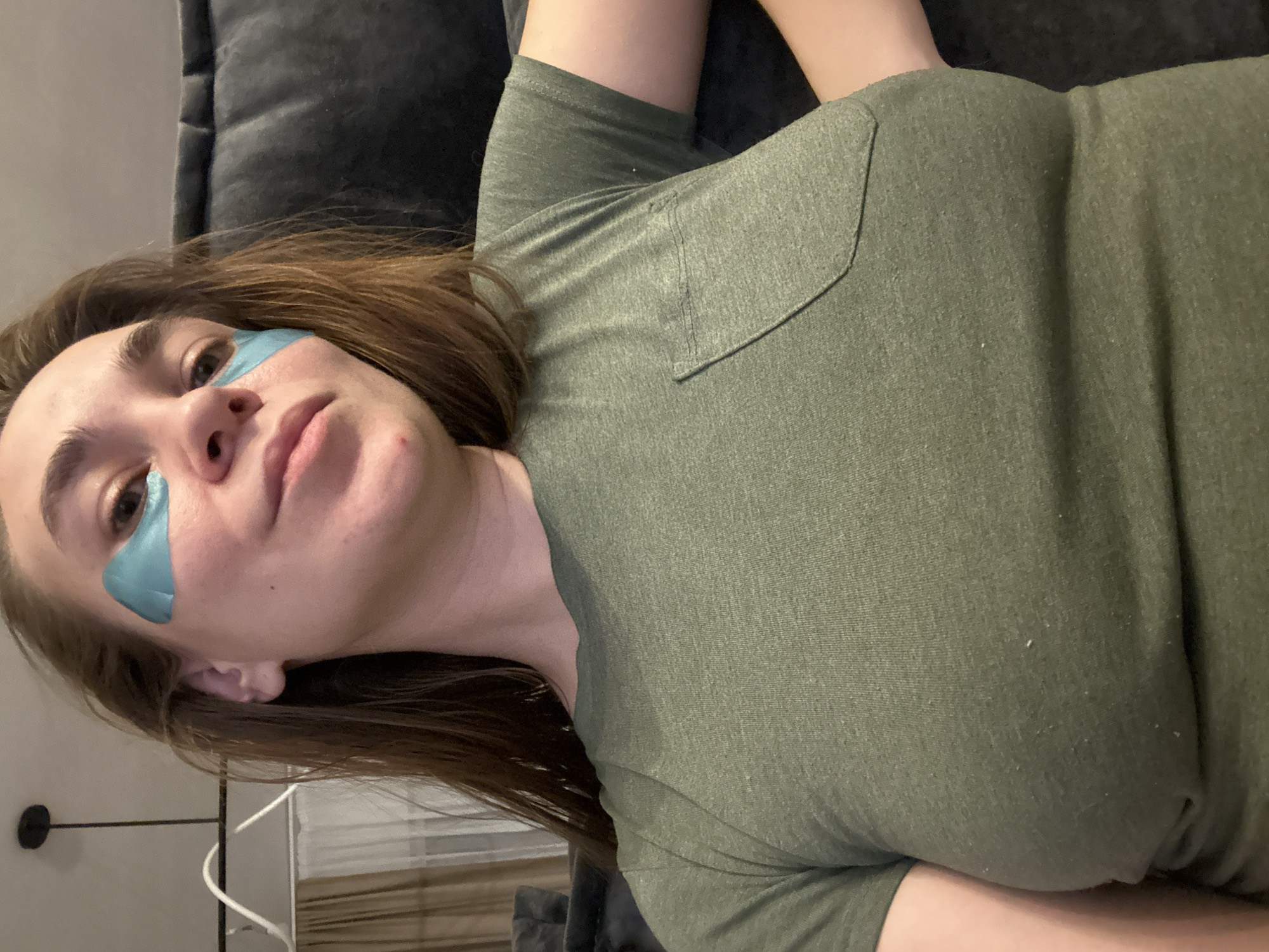 A woman is lying down on a grey surface, possibly a sofa or bed, indoors. She is wearing a short-sleeved, olive green shirt with a small pocket on the front. A light blue patch is adhered to her skin under her left eye. Her brown hair is loose. Part of a lamp and a white cable are visible in the lower left corner of the image. The location in the city of Kharnavy, Russia is not apparent from the image.