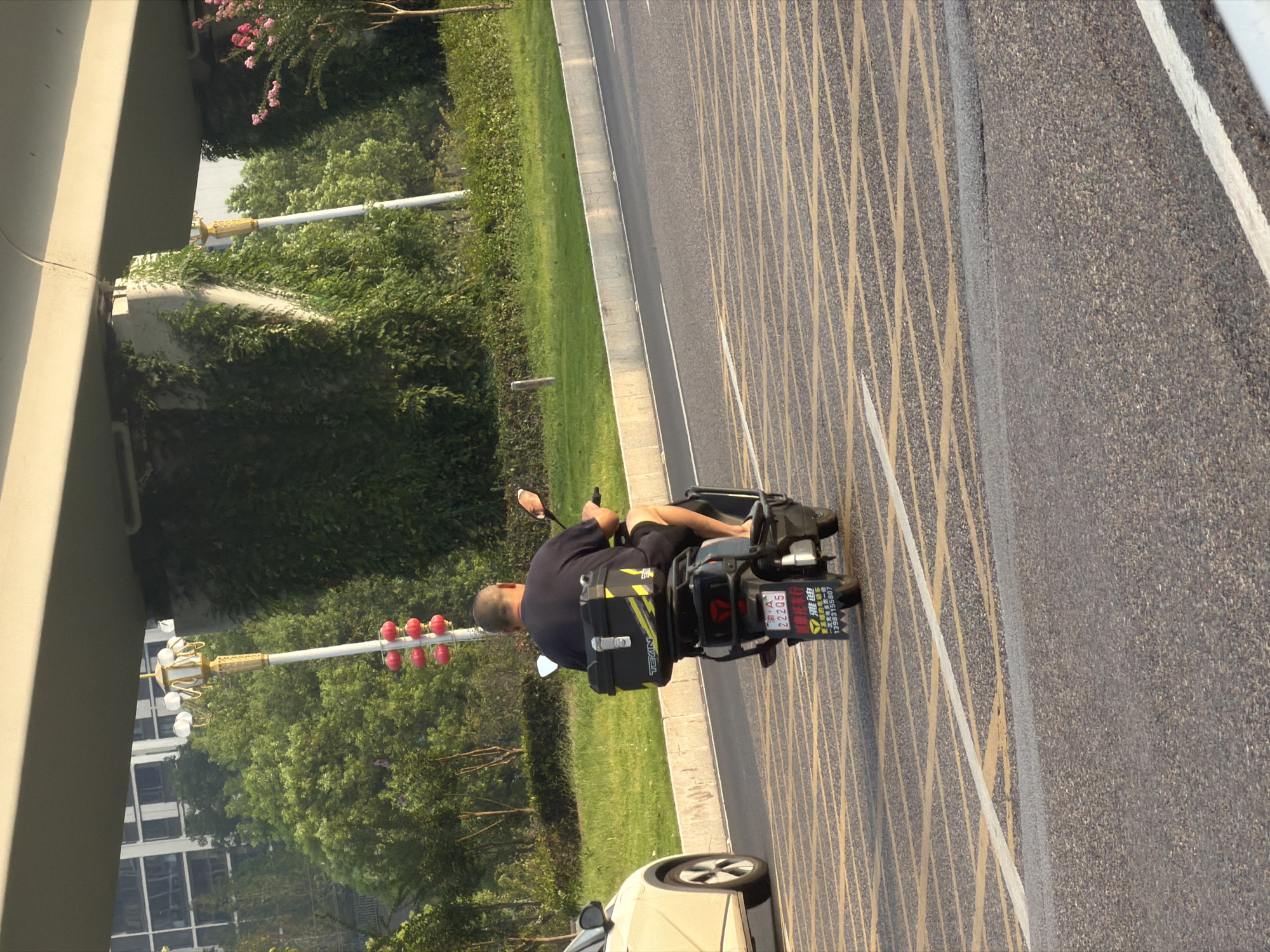 A rear view of a man riding a black electric scooter with a black storage box on the back is captured on a paved road with yellow and white lane markings. The man is wearing a dark t-shirt and dark shorts. The scooter has a license plate that reads "2220S" and some Chinese characters. A white vehicle is partially visible on the left side of the road. The background features a grassy area, lush green trees, a building with multiple windows, a street lamp with red lanterns, and a section of an elevated highway structure. The scene appears to be in the city of Jiangjin, China, based on the text on the license plate.