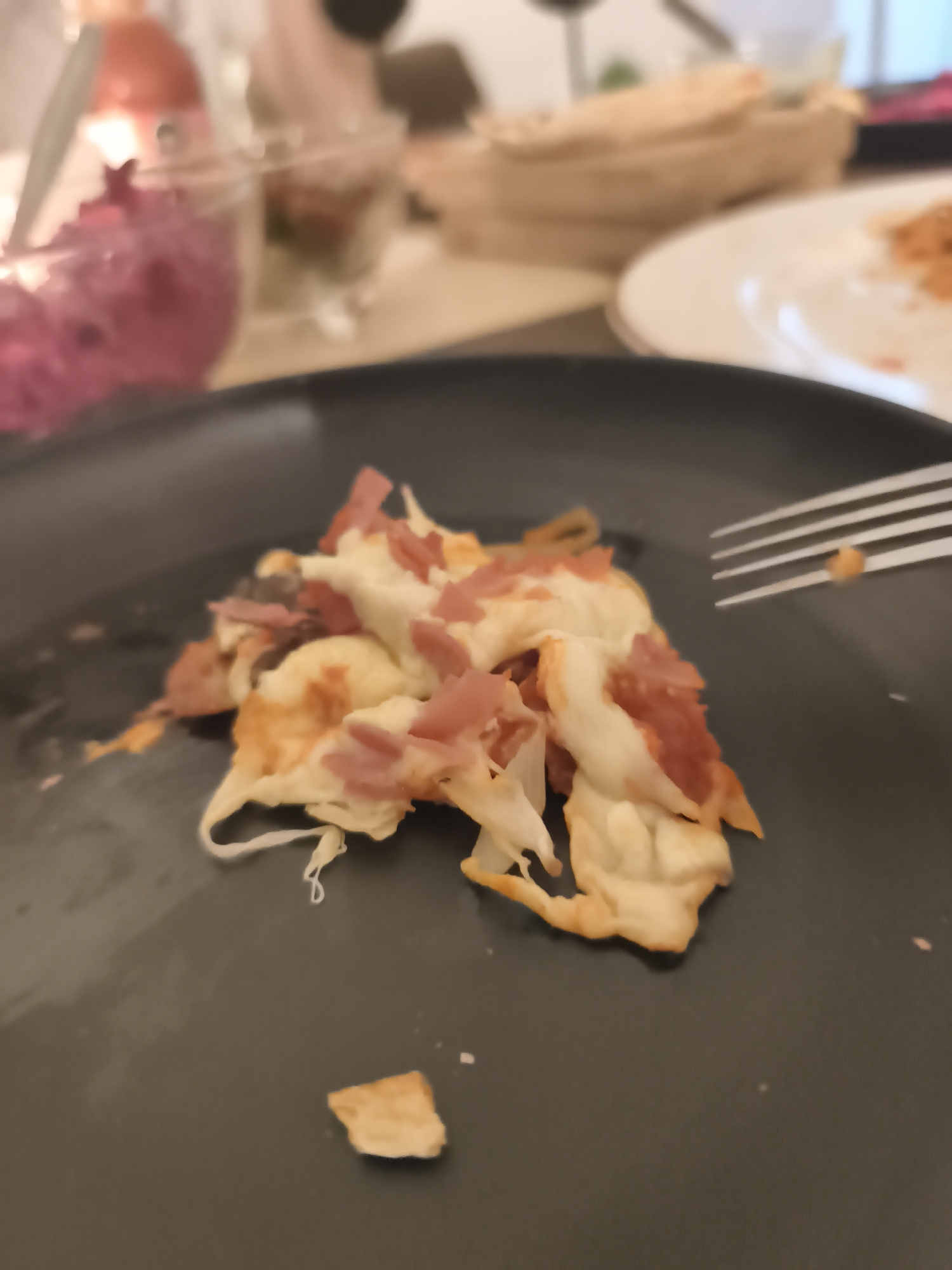 A close-up shot on a dark gray plate reveals remnants of a meal consisting of a cheese-covered food item sprinkled with diced ham. A metal fork is partially visible on the right side of the frame, suggesting the food is being or has been eaten. In the blurred background, additional plates, bowls filled with what appears to be salad or other condiments, and other table items are visible. This suggests a dining setting, likely in a restaurant or a home. The location is specified as Mexico City, Mexico.