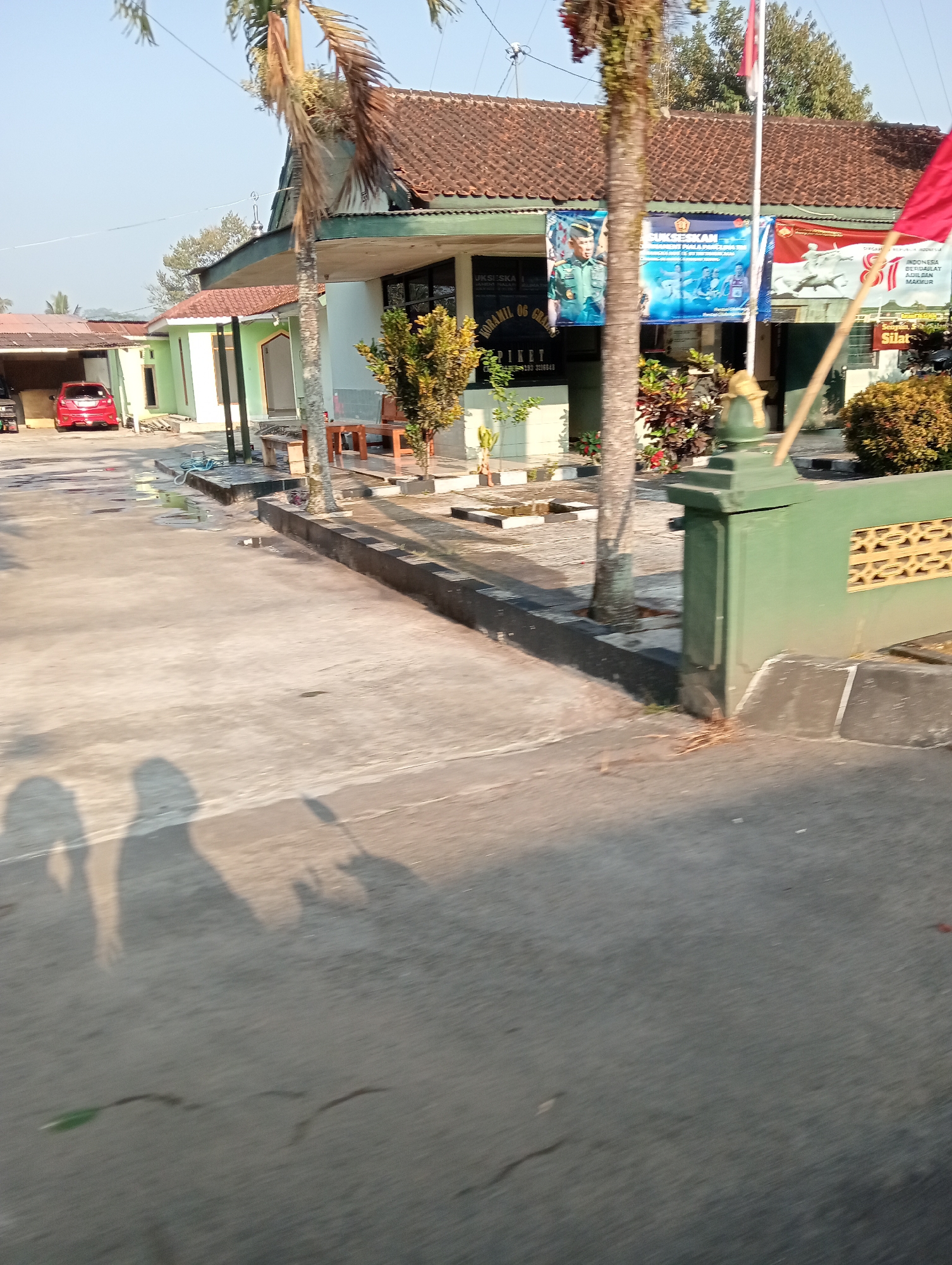 The image depicts the exterior of a building with banners displayed on its facade. The building appears to be a government or community facility, possibly a sub-district office or a village hall, located in Grabag, Indonesia. The banners are printed in Indonesian, with some featuring images of military personnel and slogans related to national pride or community initiatives. One banner prominently displays a portrait of a man in a military uniform, alongside text that appears to be a call to action or a motivational message. Another banner shows a group of people participating in a sporting event, possibly swimming. A red flag is also visible, flying from a pole near the building. The building itself has a tiled roof and a light green painted exterior. In front of the building, there is a paved area with some potted plants and outdoor furniture. A red car is parked to the left of the building. The overall scene suggests a public space on a clear day, likely in the morning or late afternoon given the shadows cast on the ground. The shadows of two people are visible in the foreground, indicating their presence at the time the photograph was taken. The environment appears to be semi-urban or rural, with palm trees and other vegetation surrounding the structure. The presence of multiple banners suggests an event or ongoing campaign being promoted at this location.
