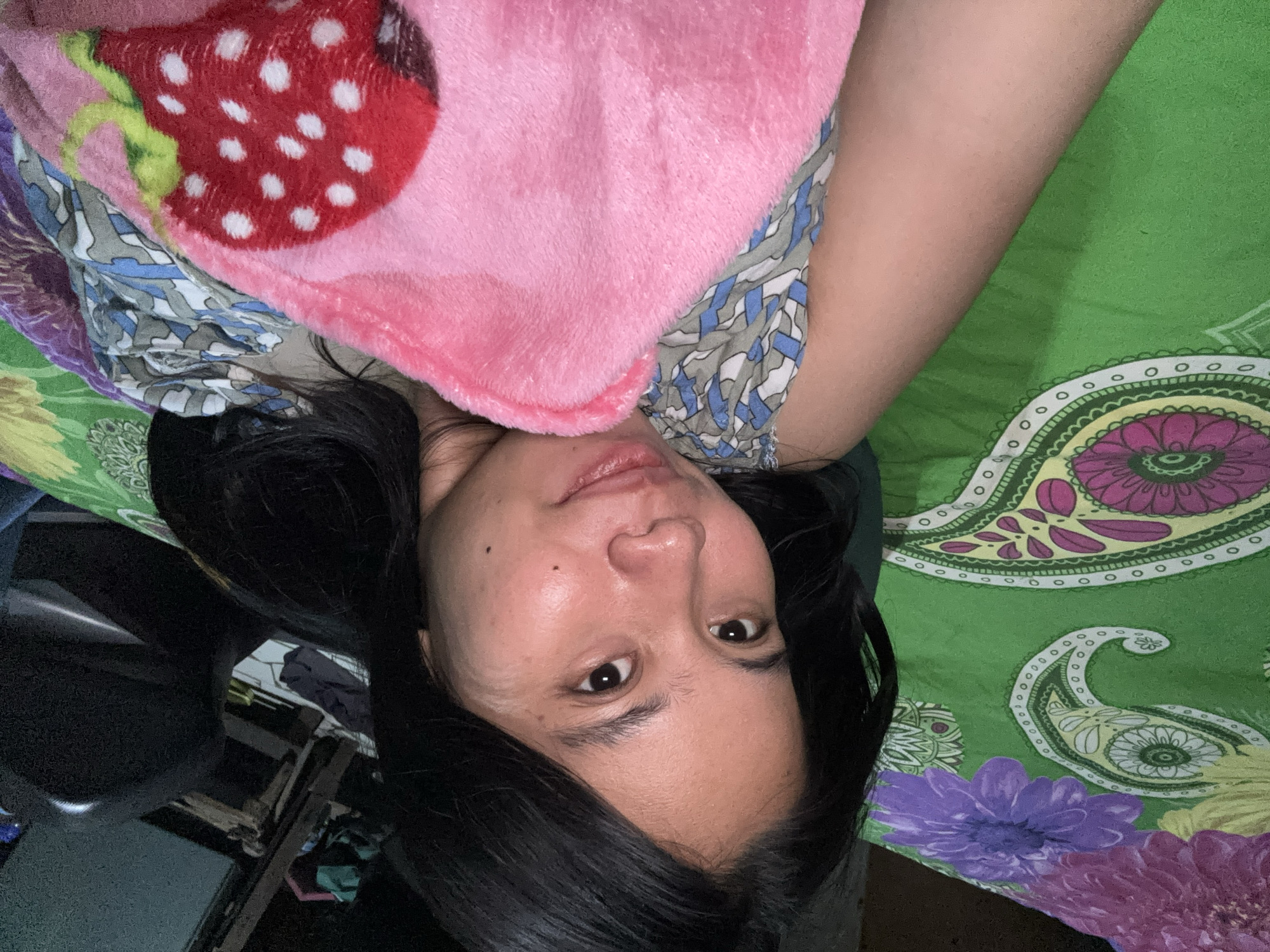 A young woman with long, dark hair lies on her side, looking towards the camera. She is covered by a pink blanket with a strawberry pattern and a patterned fabric. The background consists of a green patterned sheet with floral and paisley designs. The image appears to be taken indoors, possibly in a bedroom, with a dark object, possibly a chair, visible in the upper left corner. There are no other individuals or objects of particular interest in the frame. The lighting is dim, suggesting it might be nighttime or the room is not well-lit. The specific location in East Jakarta, Indonesia cannot be determined from the visual information provided in the image.