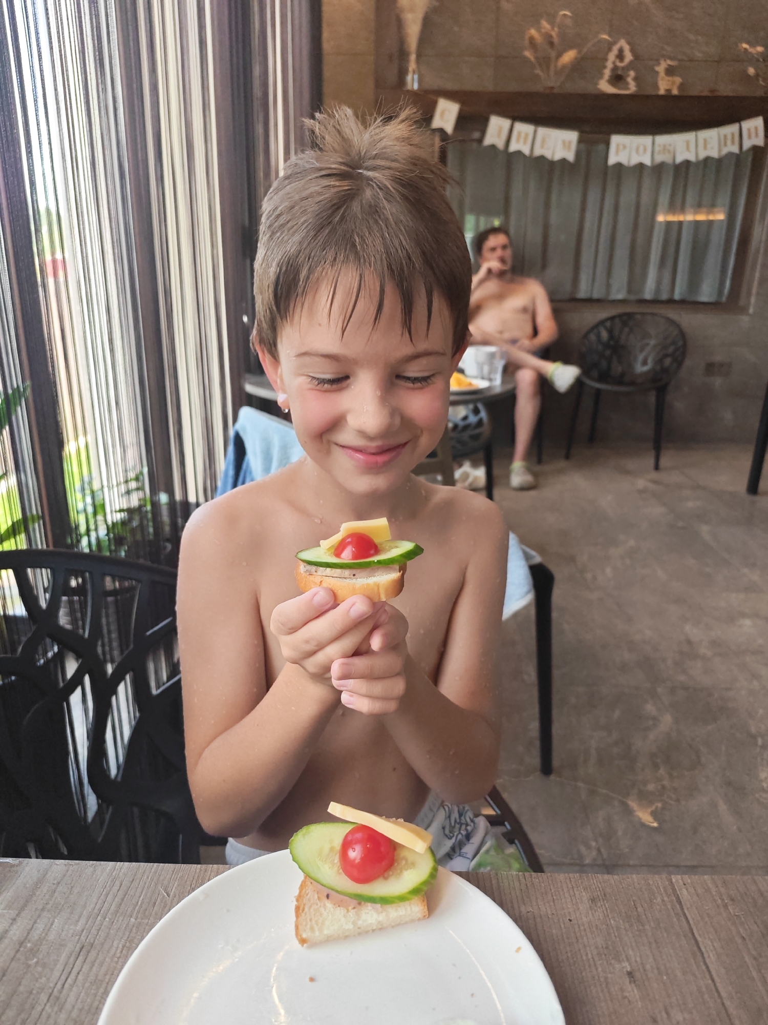 A young boy is in the foreground of the image, holding a small open-faced sandwich in his hands. The sandwich is on a slice of bread and topped with a slice of cucumber, a cherry tomato, and a piece of cheese. Another similar sandwich sits on a white plate in front of him. The boy is shirtless and appears to be wet, suggesting he might have been swimming. He has brown hair and is smiling broadly as he looks down at the sandwich.
In the background, a man is seated on a chair, also shirtless. He is looking towards the right of the frame. Behind him, a banner with Cyrillic letters hangs across a window or curtained area, reading "С ДНЕМ РОЖДЕНИЯ," which translates to "Happy Birthday." The setting appears to be outdoors, possibly near a pool or recreational area, with striped curtains on the left and a stone or concrete floor. The lighting suggests it is daytime. The overall atmosphere is cheerful and celebratory.