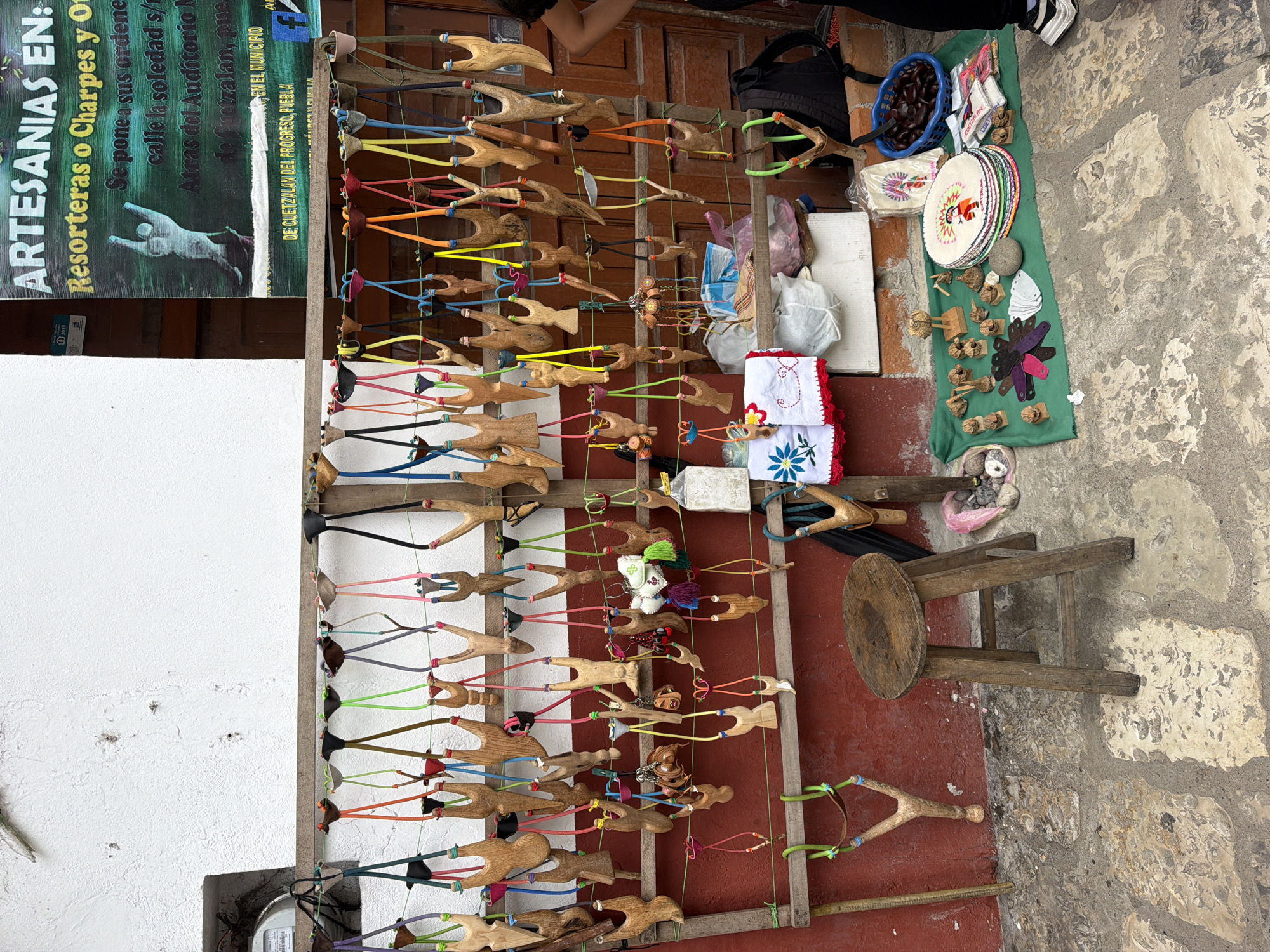 The image shows a street-side display of handcrafted items in Cuetzalan del Progreso, Puebla, Mexico. The primary items for sale are wooden slingshots, each with a unique carved handle and brightly colored rubber bands. These are hung on a wooden rack against a white wall and a red wall. Alongside the slingshots, various embroidered crafts, small figurines, and other artisanal goods are laid out on a green cloth on the stone pavement. A small wooden stool sits nearby. A sign advertising the "Artesanias" as "Resorteras o Charpes y Otros" (slingshots and other crafts) can be seen, with text indicating the location: "Calle la Soledad s/n, Atras del Auditorio Municipal." A basket of dark-colored fruit or seeds is also part of the display.