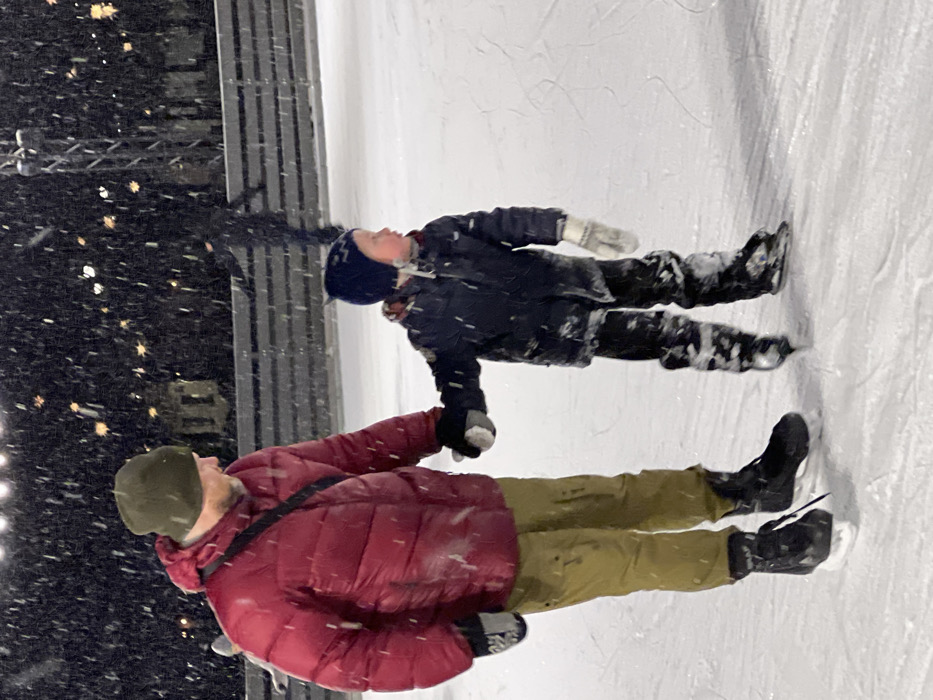 This image depicts an outdoor ice rink at night in Saint Petersburg, Russia, during heavy snowfall. An adult male and a young male child are ice skating. The adult, positioned on the left, wears an olive green beanie, a red puffed winter jacket with a black strap across his chest, olive green pants, black ice skates, and grey-white mittens; he has a beard. He holds the right hand of the young child, who is positioned to his right. The child wears a dark blue knit hat with ear flaps, a dark blue or black puffed winter jacket, dark snow-covered pants, white knit mittens, and black ice skates. Both individuals are standing on the ice, which exhibits visible skate marks. Numerous white snowflakes are suspended in the air and accumulated on their clothing. A dark, slatted fence or barrier runs horizontally across the mid-ground, separating the rink from the background. Blurred warm-toned lights, a tall dark metal structure, and distant urban architectural elements are visible beyond the barrier. A third, blurred figure on skates is discernible behind the barrier in the mid-ground right.