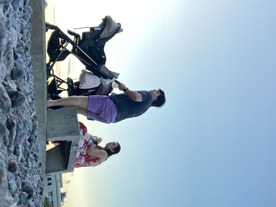 A family outing near a shoreline. A woman in a floral dress sits on a concrete structure, cradling a baby. Nearby, a man in a grey shirt and purple shorts pushes a stroller along a rocky embankment. The sky transitions from light yellow at the horizon to blue overhead. The composition focuses on the subjects against the vast sky, while the concrete and rocky terrain ground the scene.