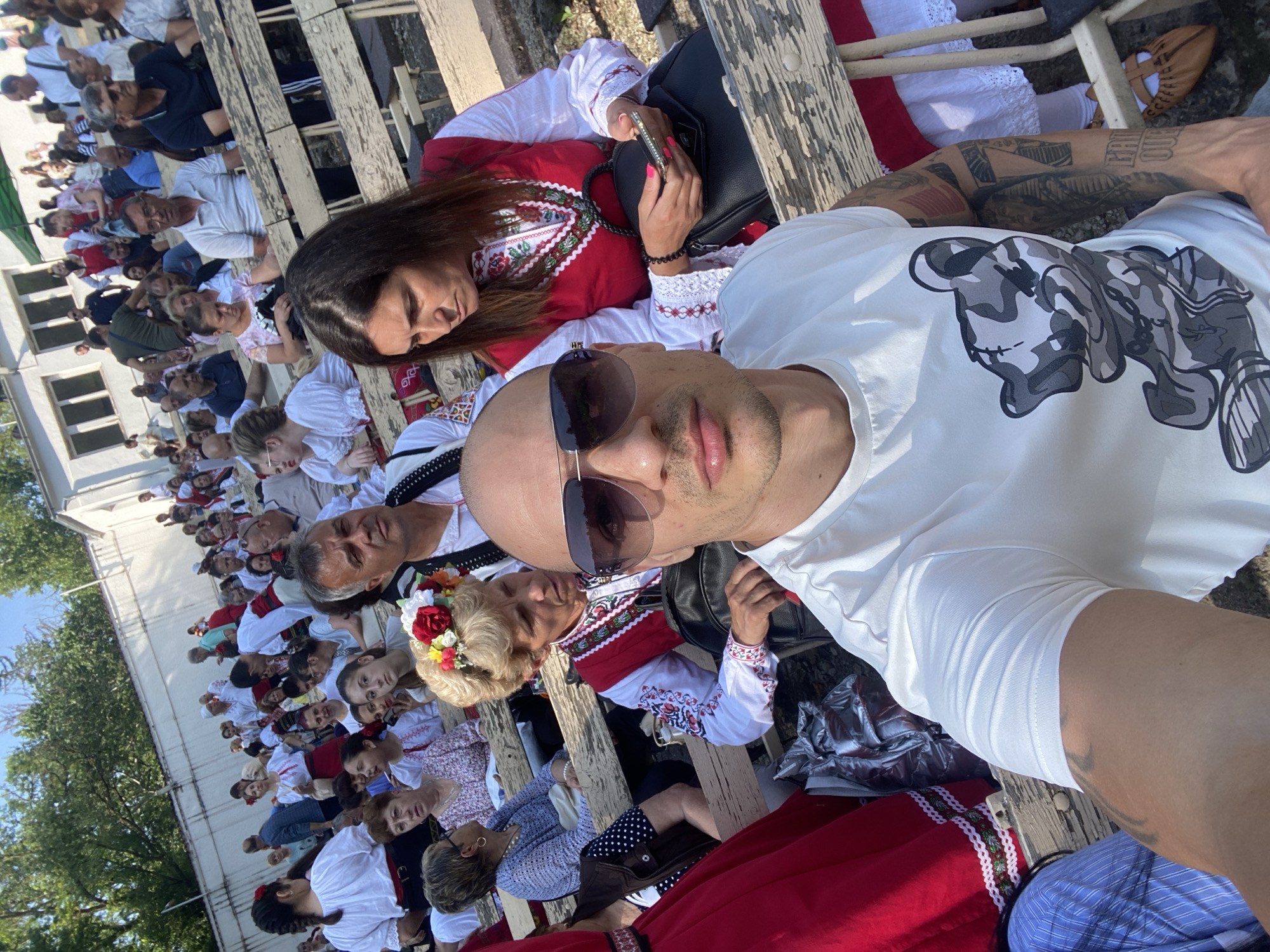 The image captures an outdoor gathering in Pleven, Bulgaria, likely a cultural event or festival, under bright daylight. In the immediate foreground, a light-skinned, bald male wearing sunglasses and a white T-shirt featuring a camouflage teddy bear graphic is taking a selfie, holding the camera at arm's length. Visible tattoos adorn his left arm.
To his right, a woman with long dark hair, dressed in a traditional Bulgarian folk costume consisting of a white embroidered blouse and a red vest, holds a smartphone with a pink case and a black handbag, looking downwards. Behind the selfie-taker, a woman with styled blonde hair and a floral wreath (red and yellow flowers) wears similar traditional Bulgarian attire, including a white embroidered blouse and a red vest, and looks slightly to her left. Adjacent to her and further back, a man with grey hair in a white embroidered shirt and a black vest, also in traditional dress, looks forward. A woman in a blue knitted top and holding a light-colored handbag is seated further back.
The midground and background reveal a large crowd of people, predominantly adults, seated on worn, tiered wooden benches. Many individuals throughout the crowd are dressed in traditional Bulgarian folk costumes, characterized by white embroidered shirts/blouses and red or black vests. In the far background, a white building with multiple windows and a long, low white wall are visible, topped by green foliage from trees under a clear blue sky. The setting appears to be an outdoor public space, possibly an amphitheater or an area with permanent bleachers, within the city of Pleven.