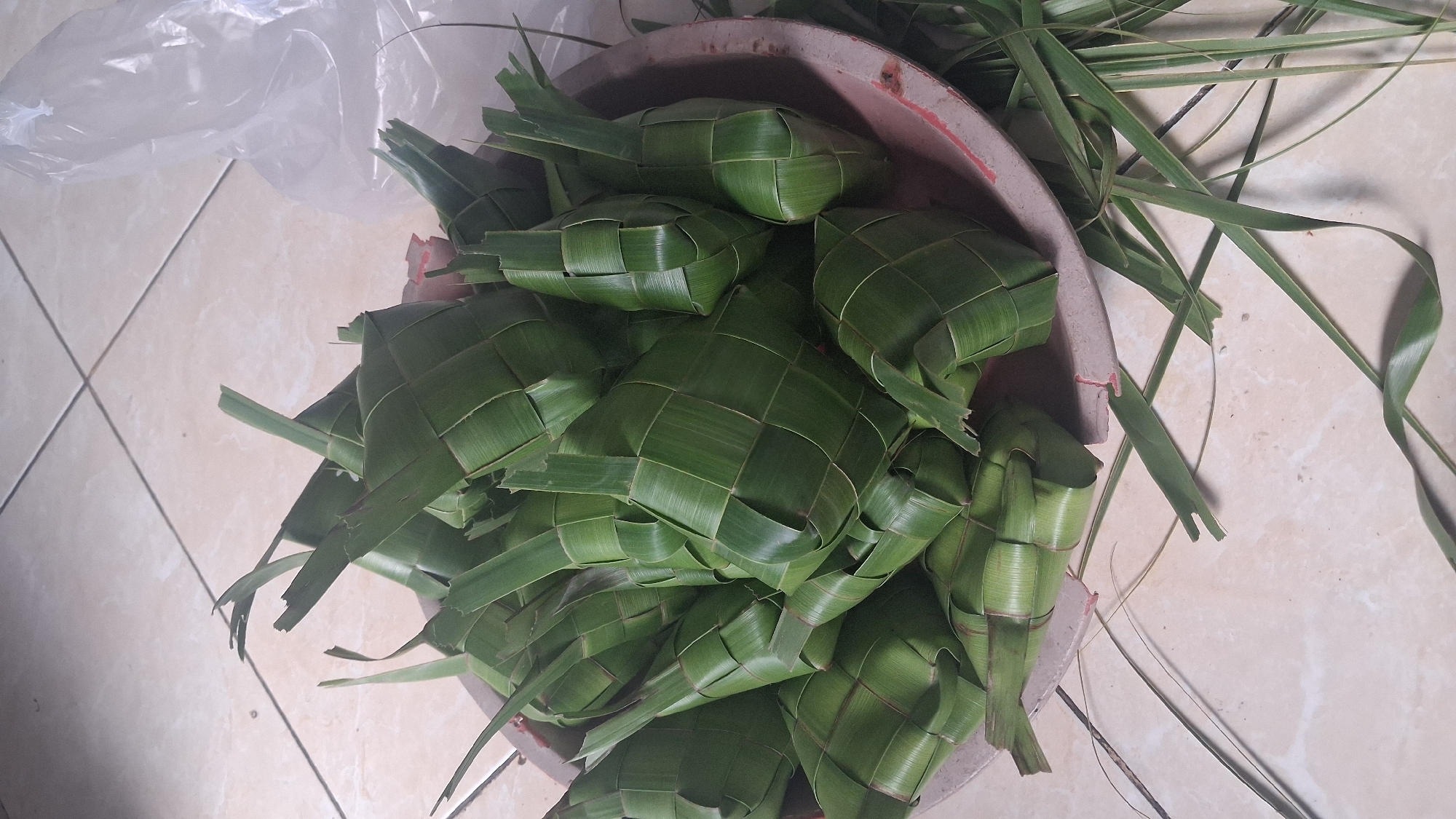 A close-up, high-angle shot shows a shallow, circular dish filled with numerous diamond-shaped parcels wrapped in green palm leaves. These parcels, known as ketupat, are a traditional rice cake wrapped in woven palm or pandan leaves, commonly eaten during festive occasions in Indonesia. The ketupat are tightly packed in the dish, with some of their pointed ends protruding upwards. Surrounding the dish, on a light-colored tiled surface, are loose green palm leaves and some dried grass. To the right of the dish, a crumpled, translucent plastic bag is partially visible. The overall scene suggests a domestic setting in Denpasar, Indonesia, likely in preparation for a meal or celebration.