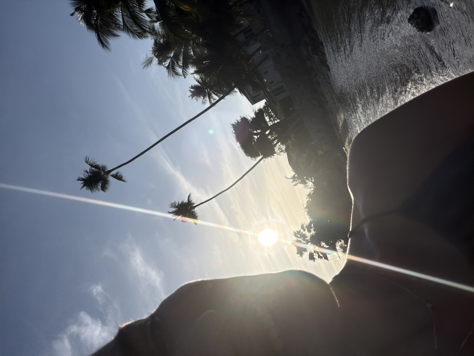 The image is a selfie taken outdoors at sunset on a beach in Ko Samui District, Thailand. The foreground is dominated by the profile of a person's head and shoulder, silhouetted against the bright sun. Palm trees are visible in the background, their fronds dark against the sky. The sun is low on the horizon, casting a warm glow and creating lens flare that streaks across the image. The sea is to the right, its surface shimmering with reflected sunlight. In the distance, a building and more palm trees are visible. The overall impression is one of a serene tropical evening.