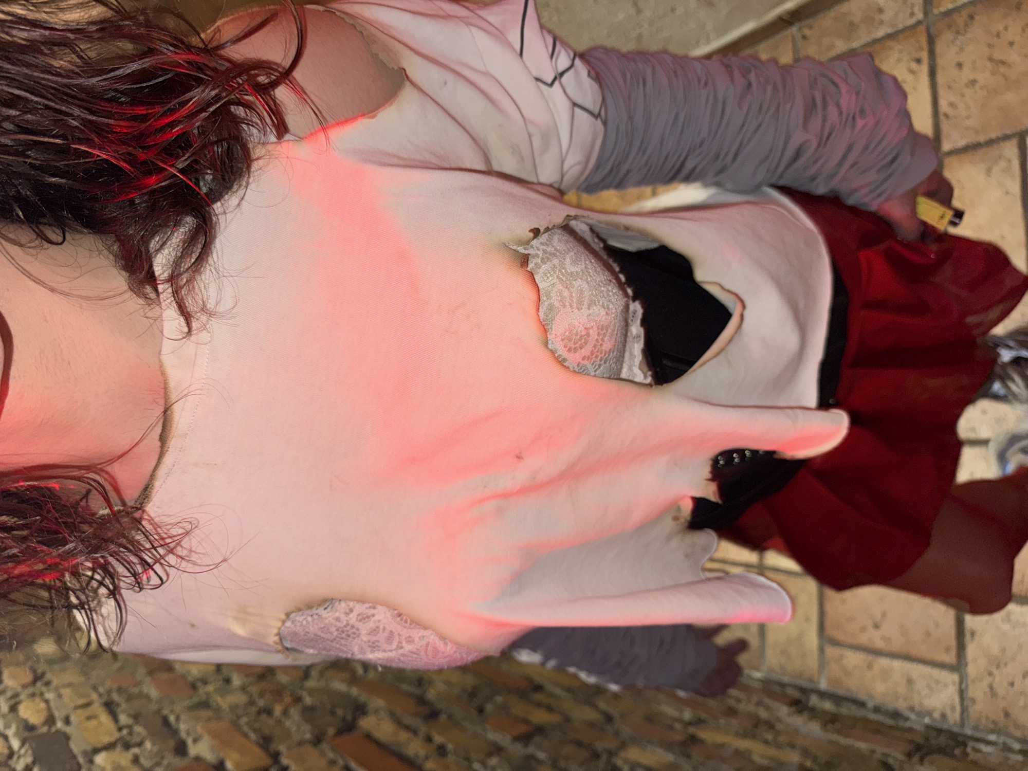 The image shows a person lying on a brick floor, possibly outdoor pavement. The person has dark hair with red highlights. They are wearing a white t-shirt with burn holes revealing a white lace bra underneath. They also wear a gray, ribbed long-sleeved shirt and a red skirt. The person holds a yellow lighter in their right hand. No evidence in the image indicates the location is Yerevan, Armenia.