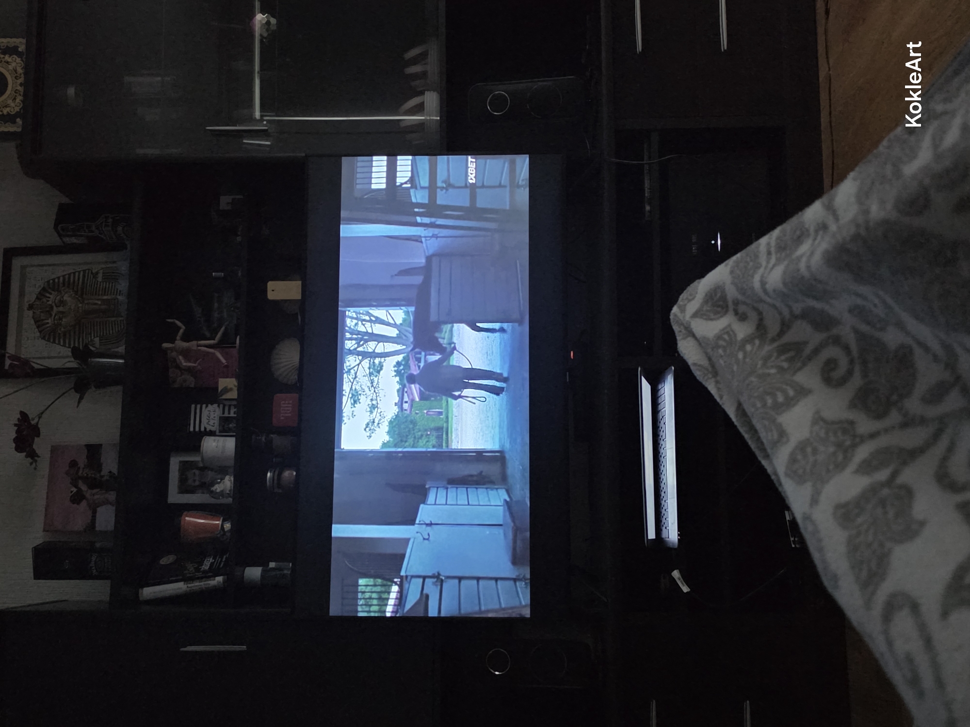 In a dimly lit living room in Melhus, Norway, a wide-screen television displays a scene from a movie or show, featuring a person walking a horse out of a stable. The television is positioned on a dark entertainment unit, which also houses a laptop and other electronic devices. The room is furnished with dark wooden cabinets and shelves, adorned with various decorative items, including a pharaoh mask, a painting, and what appear to be bottles. A patterned blanket partially covers the foreground, suggesting someone is seated or lying down watching the screen. Two black speakers are placed on either side of the television.