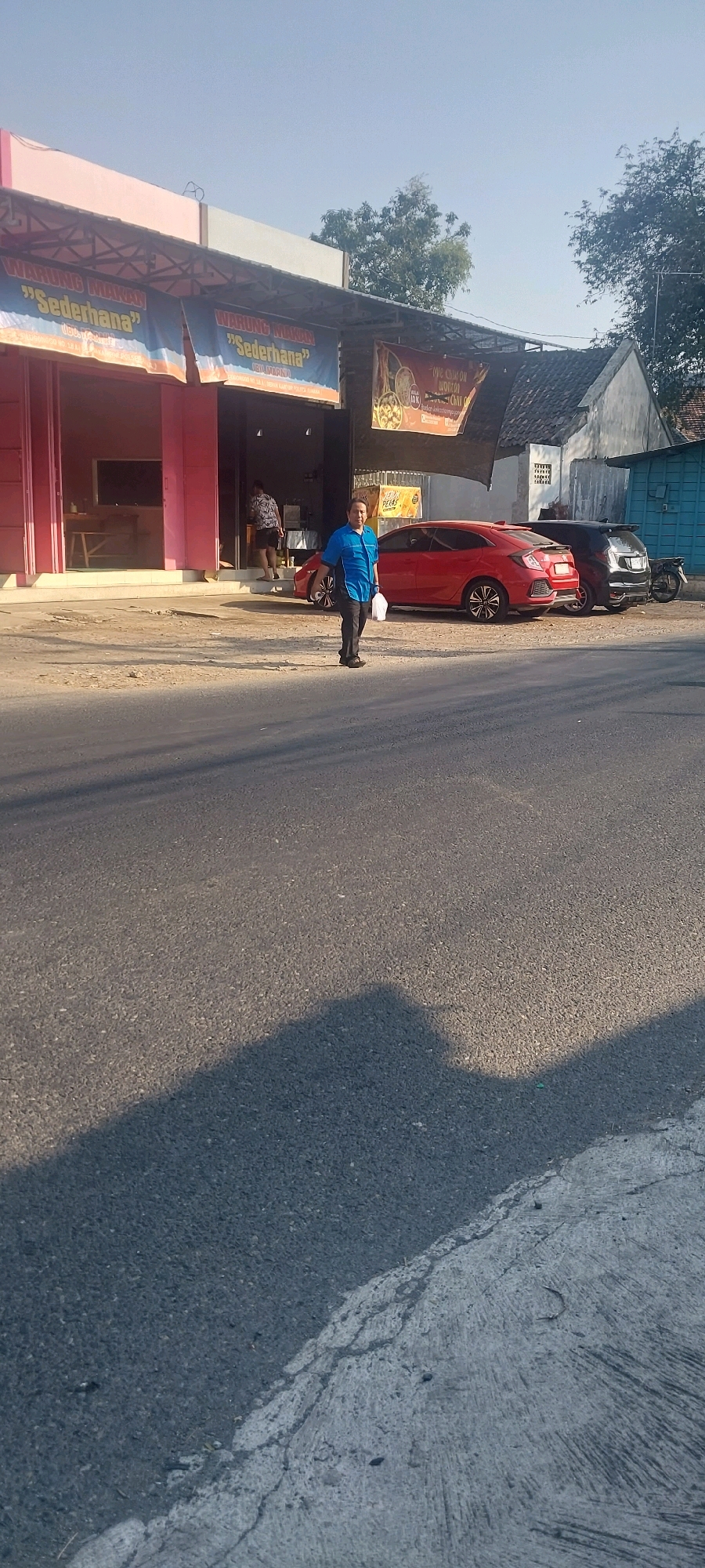 The image shows a street scene in Juwana, Indonesia. In the background, there is a building with a pink facade and a sign reading "WARUNG MAKAN SEDERHANA". A red car and a dark motorcycle are parked in front of the building. A person in a blue shirt is walking on the sidewalk, carrying a white plastic bag. Another person is visible inside the building, standing near the entrance. The foreground shows a paved road with a shadow cast on it. The sky is clear and blue.