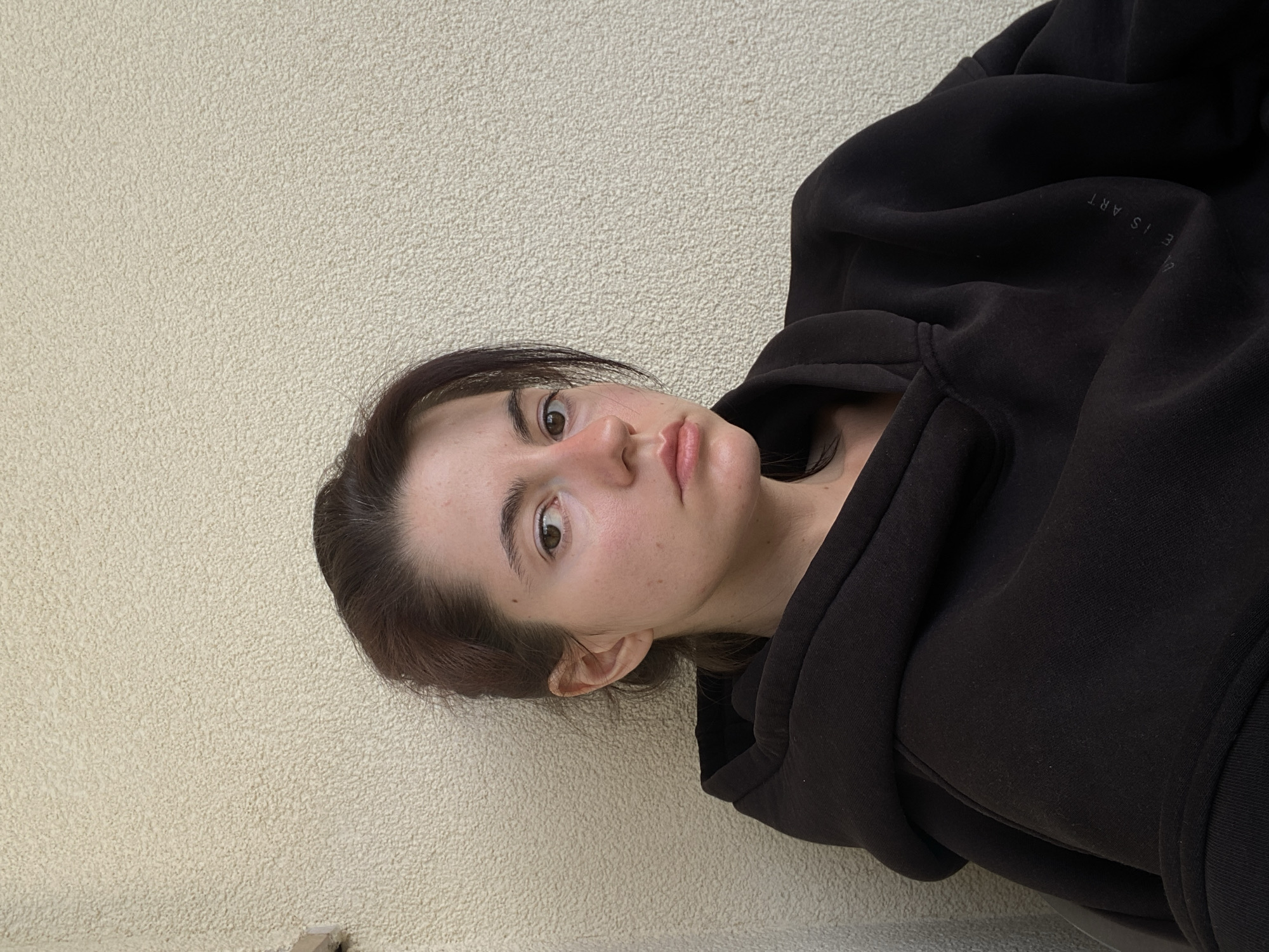 A woman is posing indoors against a light beige wall. She is wearing a dark-colored hoodie with some text visible on the right shoulder that reads "E IS ART". Her dark hair is pulled back, and she is looking directly at the camera with a neutral expression. Her face has a light complexion, and her lips are slightly pursed. The setting appears to be an interior space with textured walls. The location is unspecified in the city of Unknown, Unknown.