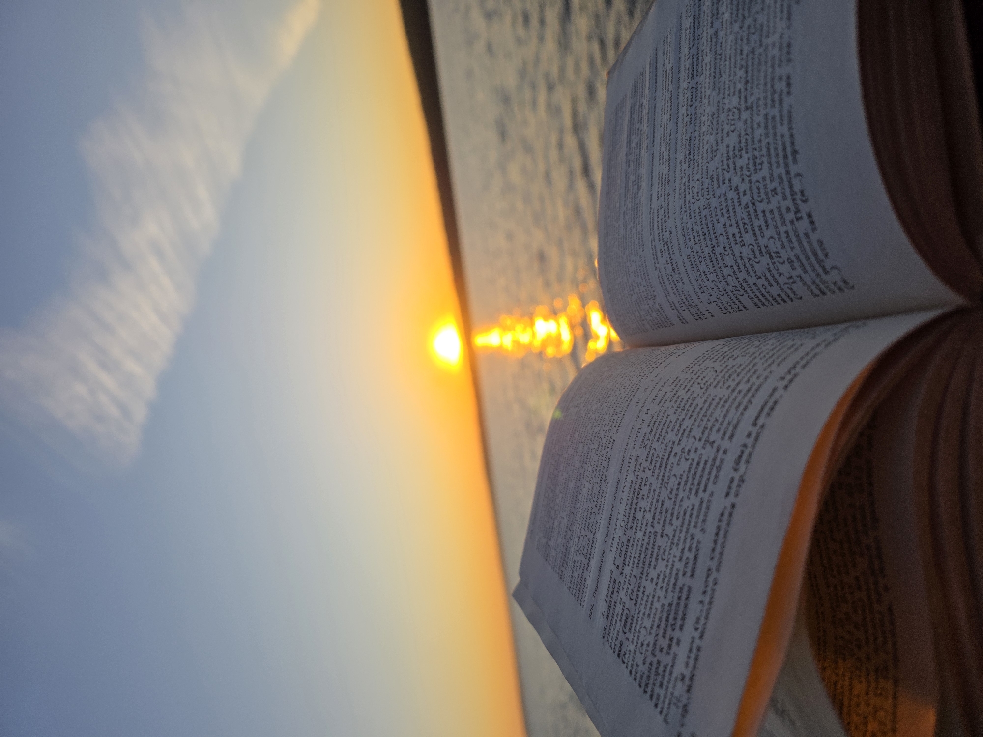 An open book lies in the foreground, its pages filled with text. The book is positioned in front of a body of water, with the sun setting on the horizon. The sky is a gradient of blue and orange, with thin clouds streaking across the upper portion. A reflection of the sun shimmers on the water's surface. The scene appears to be a serene outdoor setting, possibly a waterfront in the city of Chaikovsky, Russia, given the context provided. There are no people visible in the image.