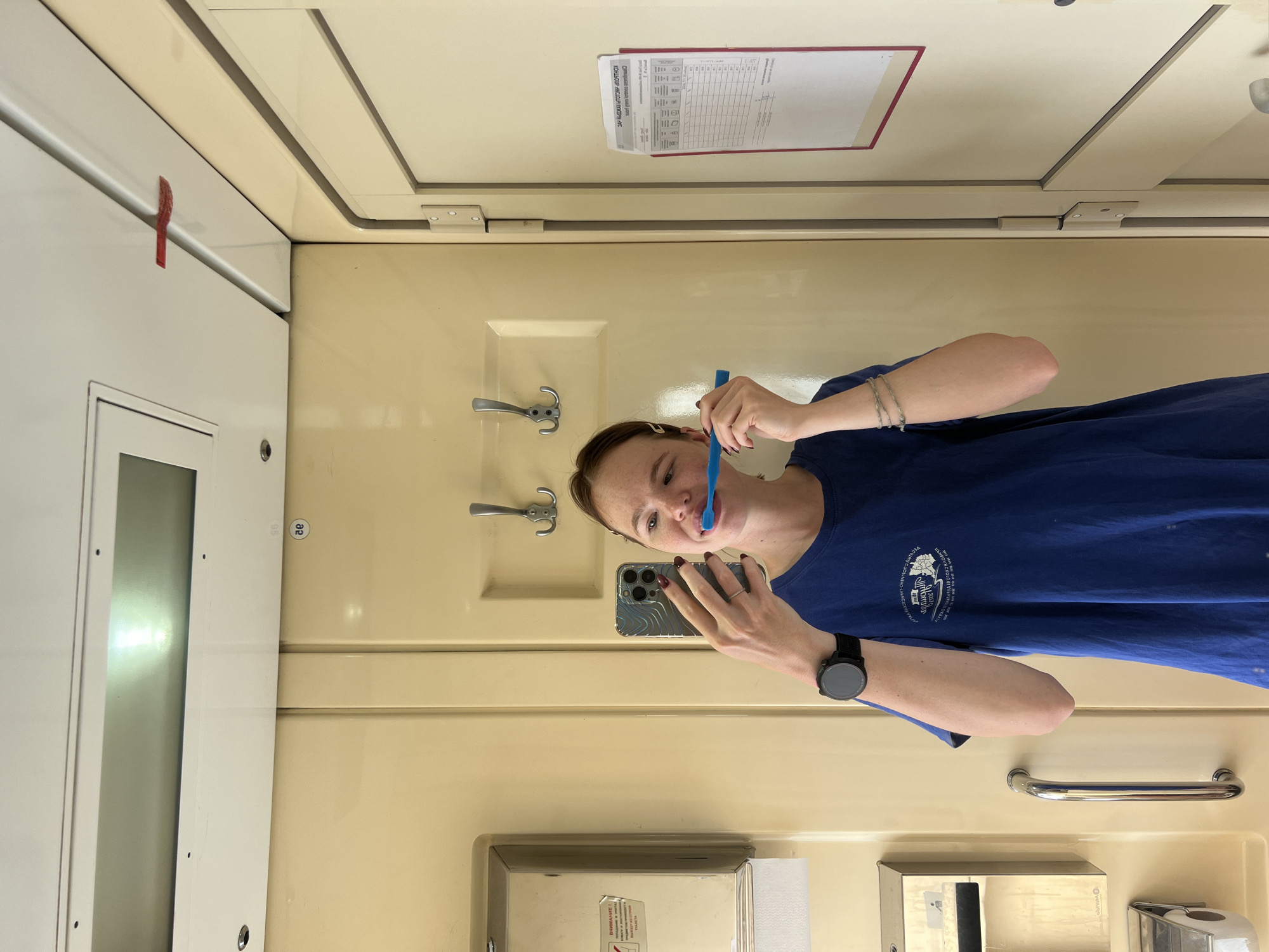 A young Caucasian woman with light skin, freckles, and light brown hair tied back is brushing her teeth in what appears to be a bathroom or lavatory compartment. She is wearing a navy blue t-shirt, a black smartwatch on her left wrist, and a silver bracelet on her right wrist. She holds a blue toothbrush in her right hand and a smartphone in her left hand, using it to take a selfie. The background consists of cream-colored paneling with hooks on the wall and a door with a red-bordered document attached to it. A metal dispenser is mounted on the wall to the left of the woman. The environment suggests an interior space within a train compartment, given the compact layout and typical amenities. The specific location in Kanash, Russia, cannot be determined from the visual information alone.