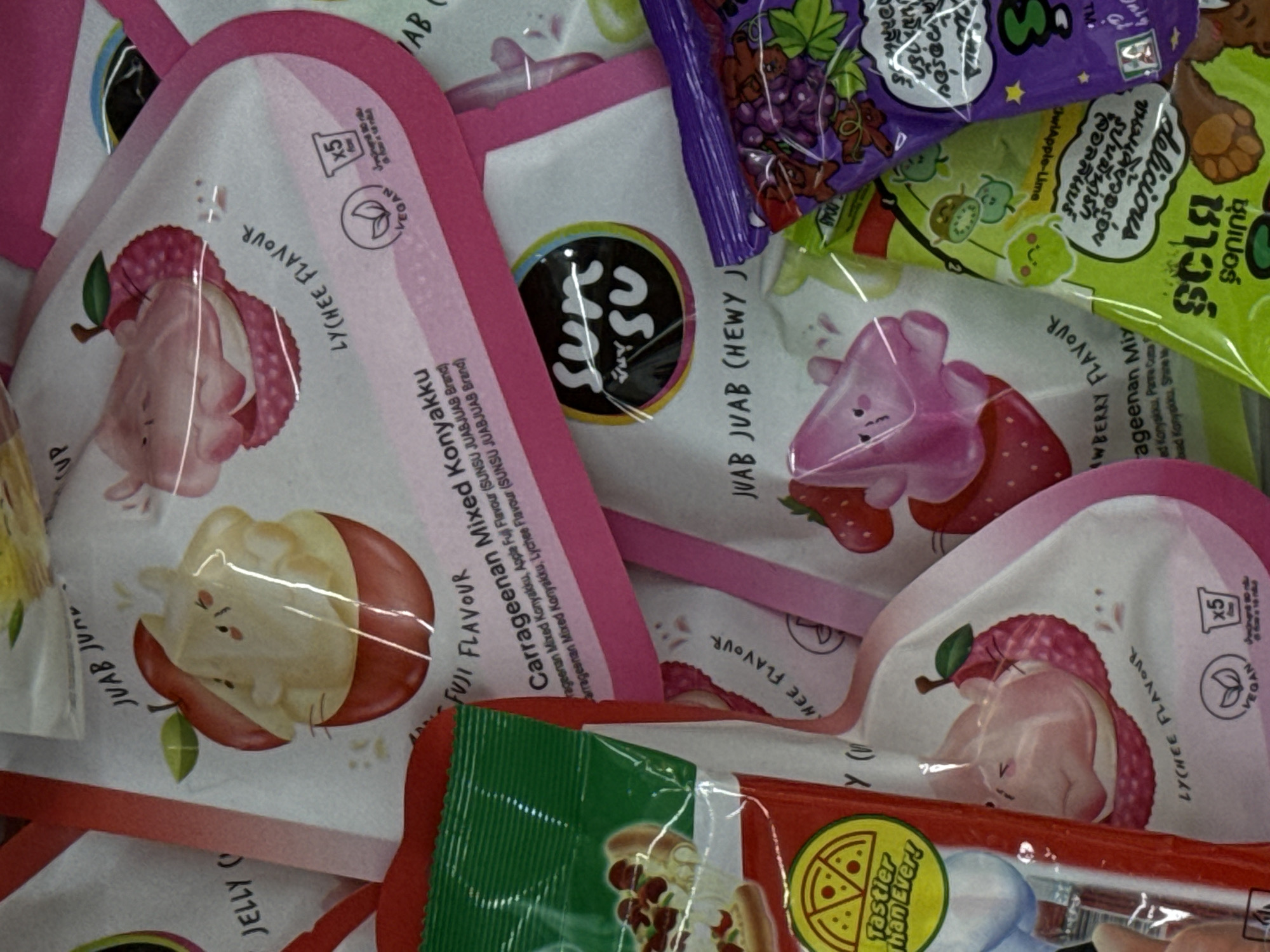 The image shows a close-up of a pile of colorful candy wrappers. The wrappers feature cartoon illustrations of fruit and characters, with text in English and what appears to be Thai. Some of the flavors advertised include Lychee, Apple Fuji, and Strawberry. The candies are jelly or gummy-like. The lighting suggests an indoor setting, possibly a store or market. There are no people or specific environmental details visible that would indicate a location in Karon, Thailand.