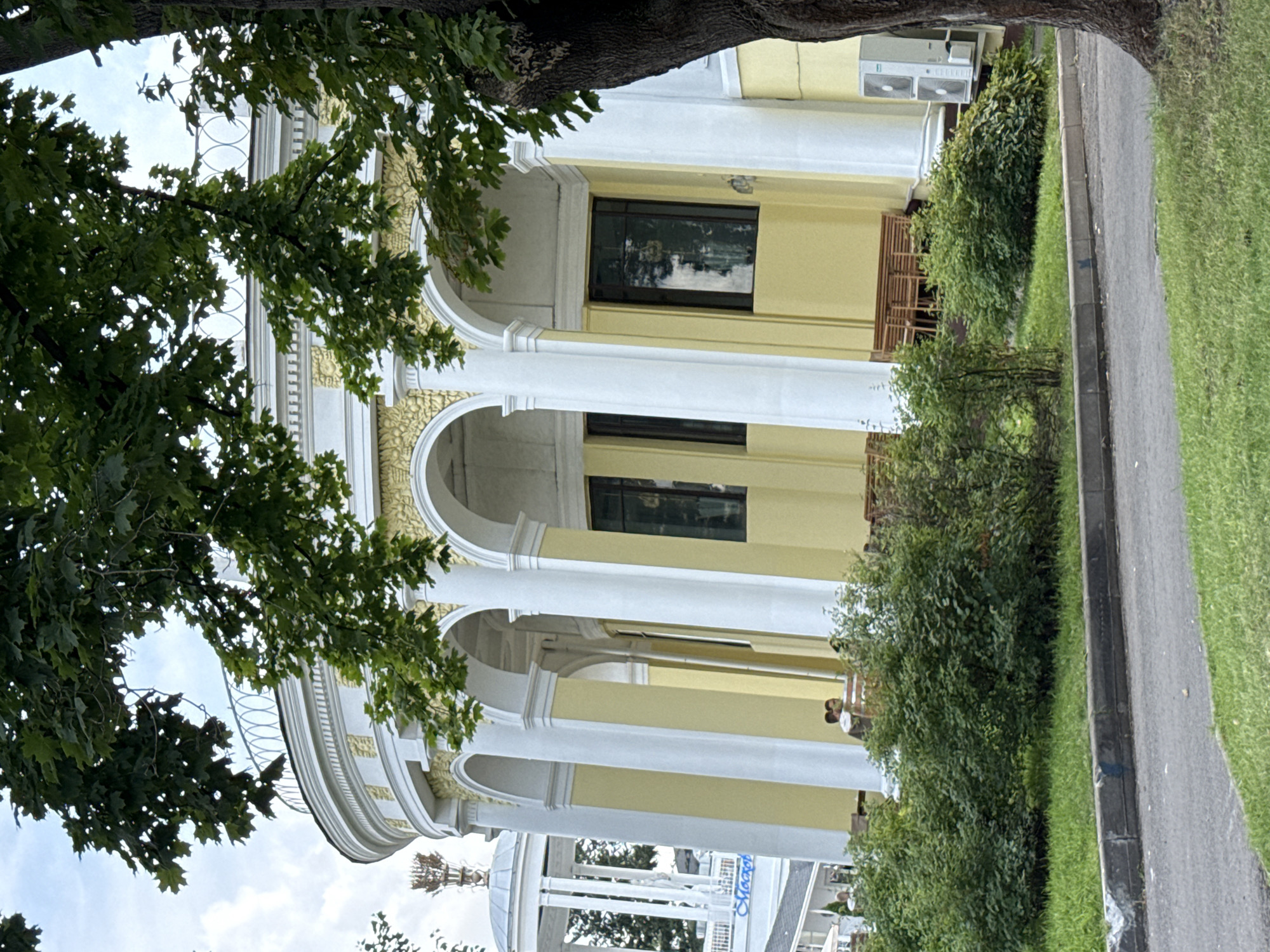 The image depicts the facade of a semicircular building with classical architectural elements, likely located in Moscow, Russia, given the presence of what appears to be the "Moscow" inscription on a white dome in the background. The building features a white colonnade with arched openings, topped by a yellow frieze and a white balustrade. Green foliage from trees hangs over the top of the structure. In front of the building, a patch of green grass is visible, bordered by a concrete curb and a paved area. Behind the bushes, there are wooden benches and two individuals sitting. Air conditioning units are mounted on the exterior wall of the building on the right side. The sky above is partly cloudy.
