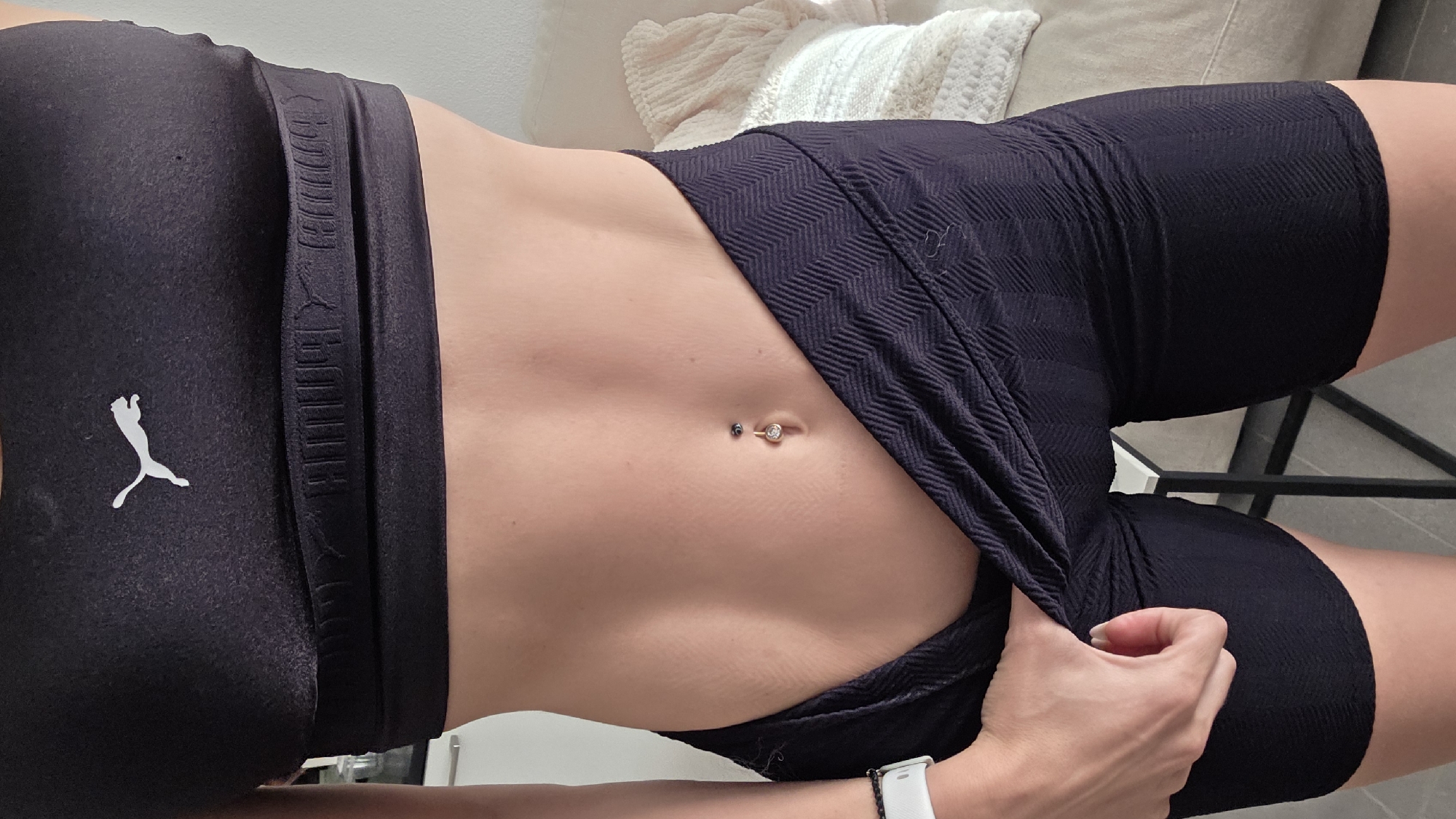 The image shows a close-up, horizontal shot of a person's torso, focusing on their midriff and upper thighs. The person is wearing black athletic wear, including a sports bra and high-waisted shorts. The sports bra features a white Puma logo on the left side, and the shorts have a subtle chevron pattern and a waistband with embossed Puma branding. A hand with neatly kept fingernails is pulling slightly at the fabric of the shorts on the right side of the image.
The person has a navel piercing with a ring that has a clear stone. The skin appears smooth and toned. The background is softly blurred, showing what appears to be a light-colored textured blanket or throw on a piece of furniture, possibly a sofa or bed.
The lighting suggests it is daytime, possibly indoors with natural light. There are no overt cues about the weather. The setting seems to be a domestic or personal space, given the soft furnishings. The overall impression is one of fitness and personal presentation. No text other than the brand logo is clearly visible.