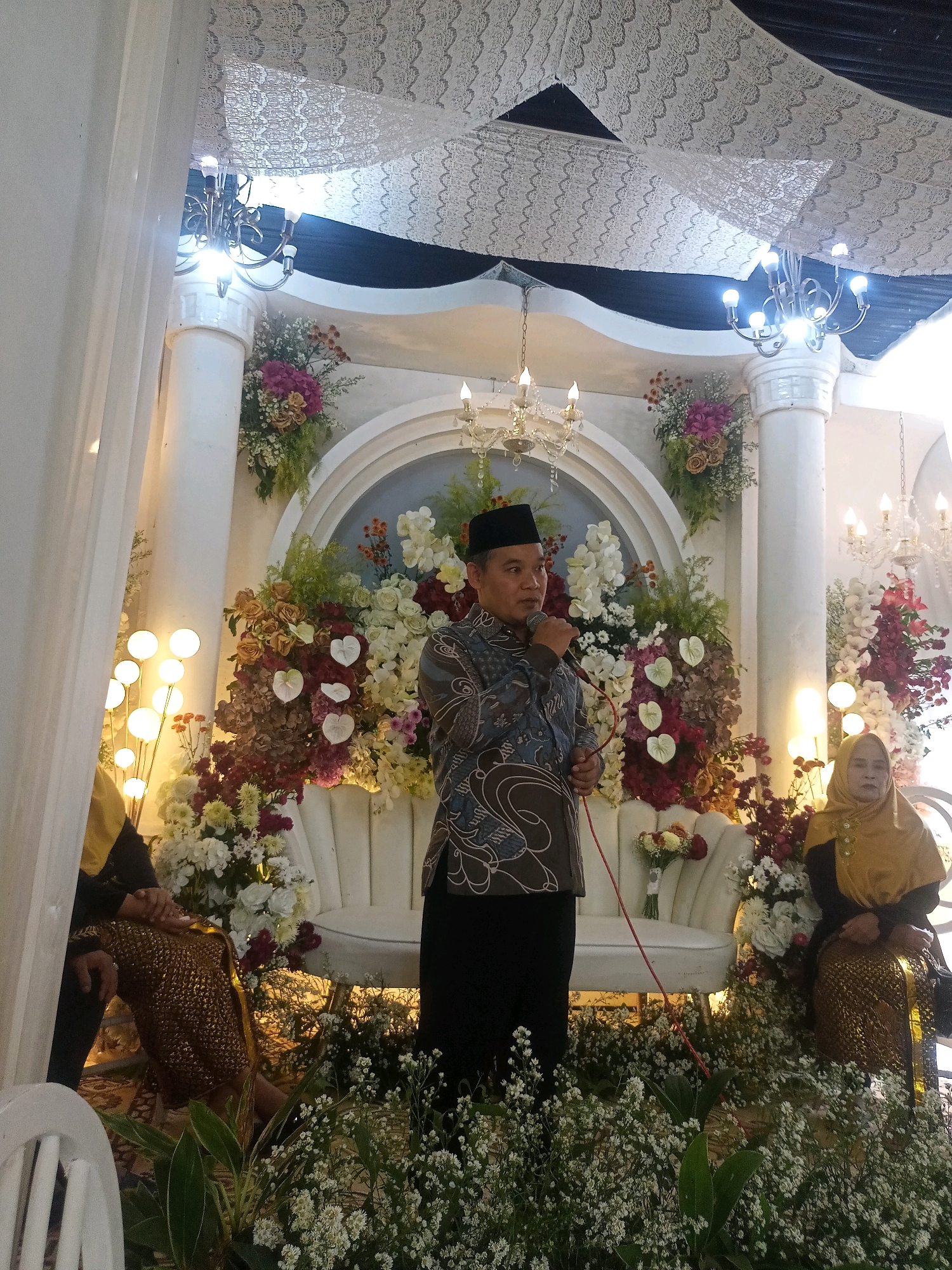 The image depicts a scene at an indoor event, possibly a wedding reception, in the city of Badamita, Indonesia. A man in a black peci hat and a patterned long-sleeved shirt stands center, holding a microphone and speaking. He is positioned in front of a white, ornate couch adorned with a bouquet of flowers. To his left, partially visible, is a person in a gold garment. To the man's right, a woman in a gold hijab and a matching patterned skirt sits on the couch. The background is elaborately decorated with white drapery, columns, floral arrangements featuring various colors of roses, orchids, and other blossoms, and chandeliers. The foreground is filled with dense white baby's breath flowers. The overall ambiance suggests a formal celebration.