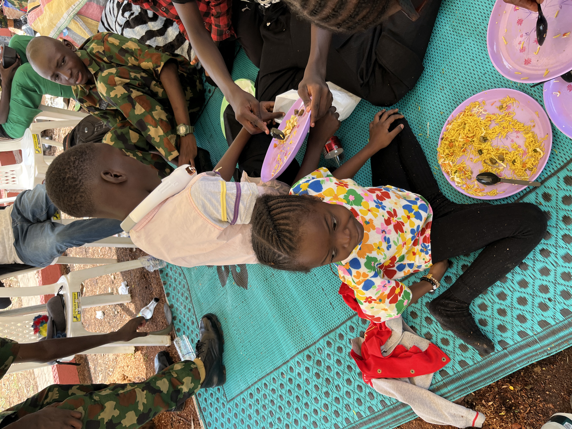 In Bar Jirgi Summa, Nigeria, a group of individuals are seated on a teal-colored mat, some in military camouflage uniforms. A young boy in camouflage is focused on a plate of rice. Nearby, a young girl with braided hair wears a colorful floral shirt and black leggings. She appears to be looking down at something on her lap, which includes a red and grey jacket. Plates of food, primarily rice dishes, are visible on the mat, with black spoons placed on them. A person in a light pink shirt and white headphones is also present. The scene suggests a communal meal in an outdoor setting, possibly a gathering or event. The ground around the mat is covered in brown dirt and debris.