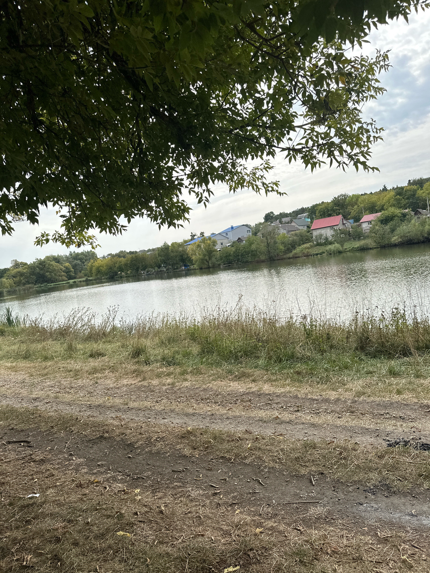 This image depicts a serene waterside scene in Makiv, Ukraine. In the foreground, a rough dirt path is visible, covered with dry grass and scattered leaves. Beyond the path, a bank of dry, overgrown grass leads down to a calm body of water. The water's surface reflects the overcast sky and the surrounding greenery. Across the water, a picturesque village is nestled on a gentle hillside. Houses with red, gray, and blue roofs are interspersed with lush green trees. The overall atmosphere is tranquil and rural, suggesting a peaceful day in the Ukrainian countryside.