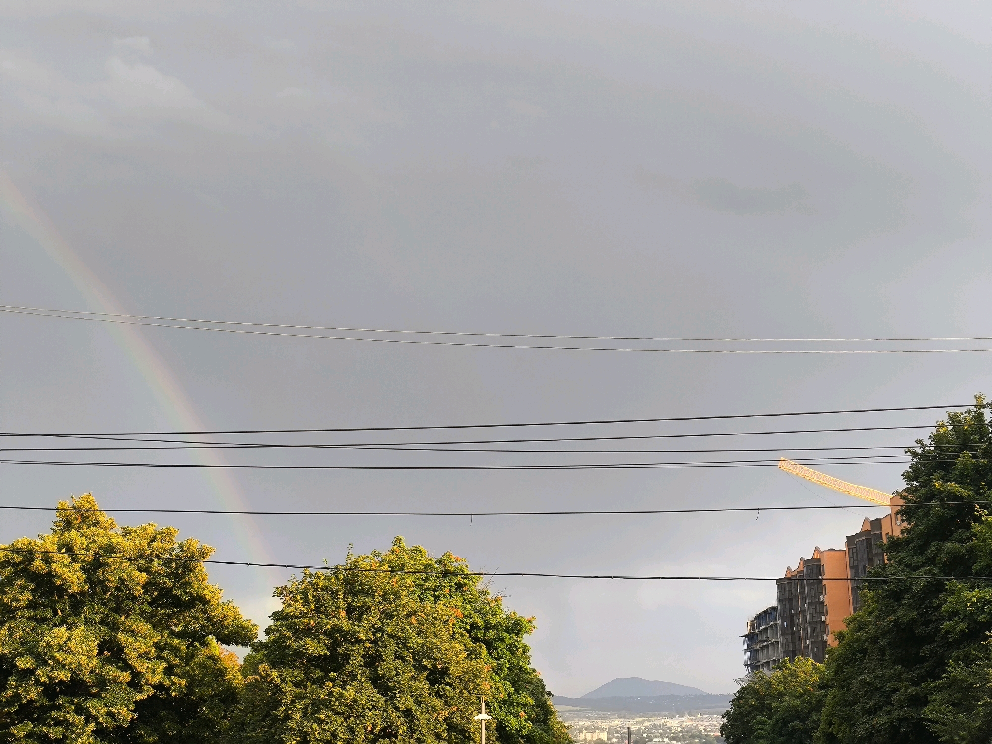 A view from a window in Yessentuki, Russia shows a grey sky with a faint rainbow arc on the left. Multiple power lines are strung across the frame. Below the sky, there are trees with yellow and green foliage. To the right, an unfinished building with orange and dark grey sections is under construction, with a yellow construction crane visible above it. In the distance, mountains are visible.