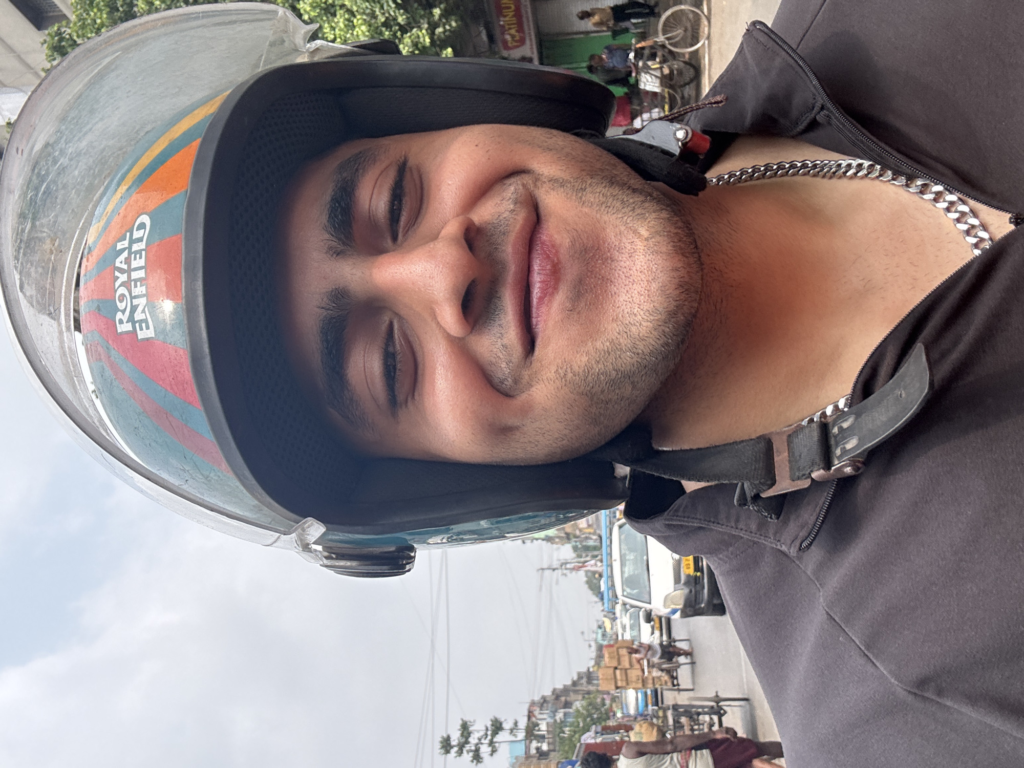 A medium-shot portrait shows a man wearing a motorcycle helmet with a clear visor and "ROYAL ENFIELD" printed on the side. The helmet features a black border and colorful stripes in orange, blue, and red. The man is wearing a dark-colored zipped-up jacket, with a silver chain visible around his neck. The background includes a street scene, likely in Kolkata, India, with buildings, traffic, and pedestrians. There are power lines overhead. The overall composition suggests the man is either sitting on a motorcycle or has just finished riding.