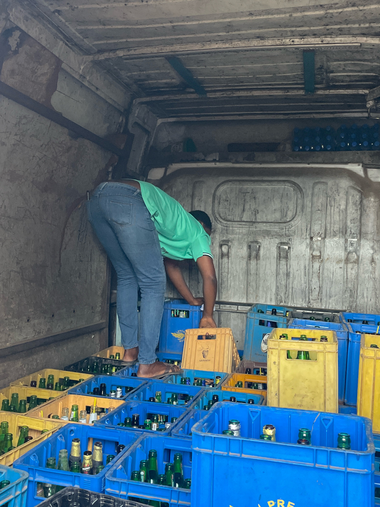 The image shows the interior of a vehicle, likely a delivery truck, filled with crates of bottled beverages. A person, wearing blue jeans and a green t-shirt, is bent over, interacting with the crates. The crates are predominantly blue and yellow, and some contain green glass bottles, possibly beer. The environment appears to be a loading or unloading area, given the presence of stacked crates. While the image itself does not explicitly mention Jos, Nigeria, it depicts a common scene of beverage distribution in urban settings.