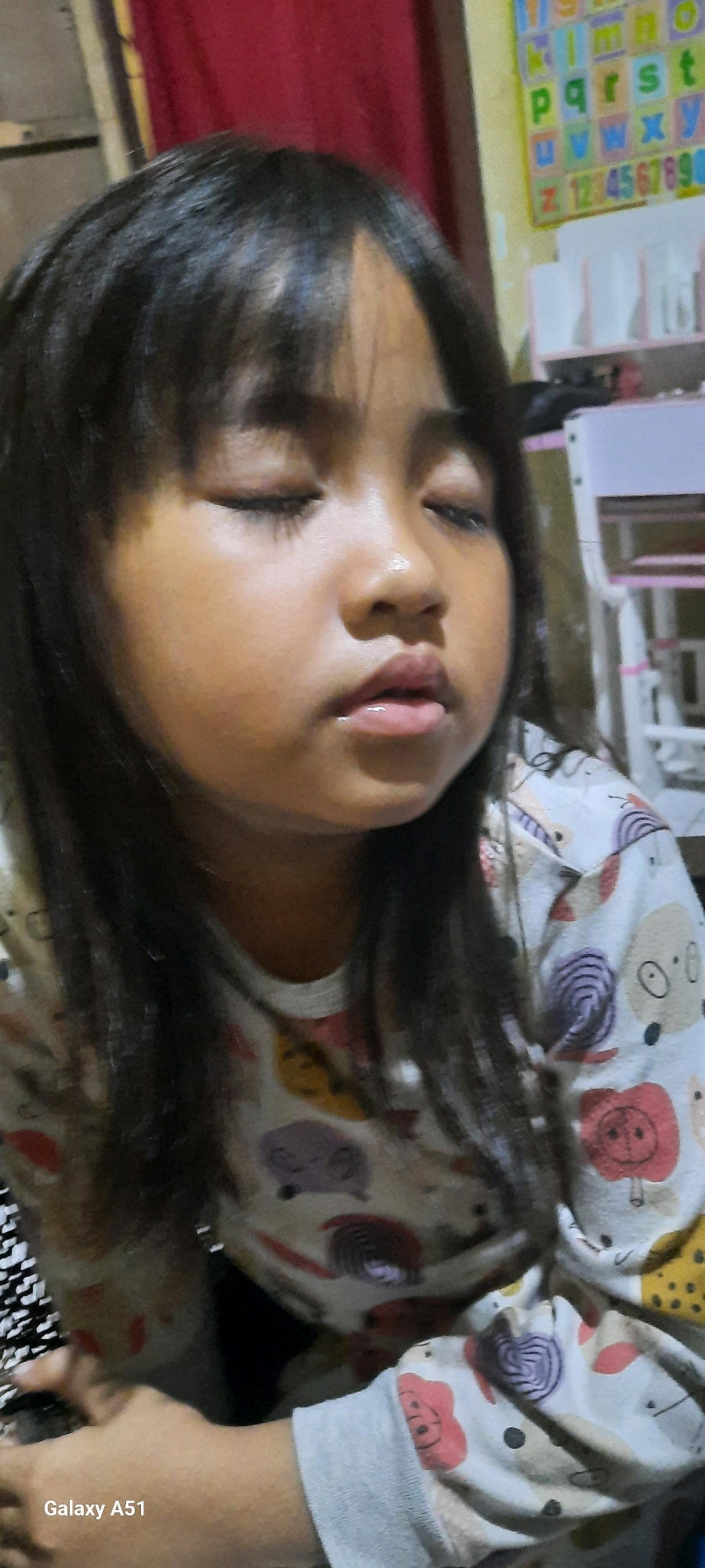 A close-up, medium-shot, eye-level, portrait of a young girl with dark hair and closed eyes is presented. She is wearing a light-colored shirt with a pattern of cartoon-like animal faces. The background is out of focus, but appears to be an indoor setting with a red curtain on the left and a colorful educational chart with letters and numbers on the right, suggesting a child's room or play area. The lighting is soft, casting a gentle glow on her face. The image is geotagged to Jatitengah, Indonesia.