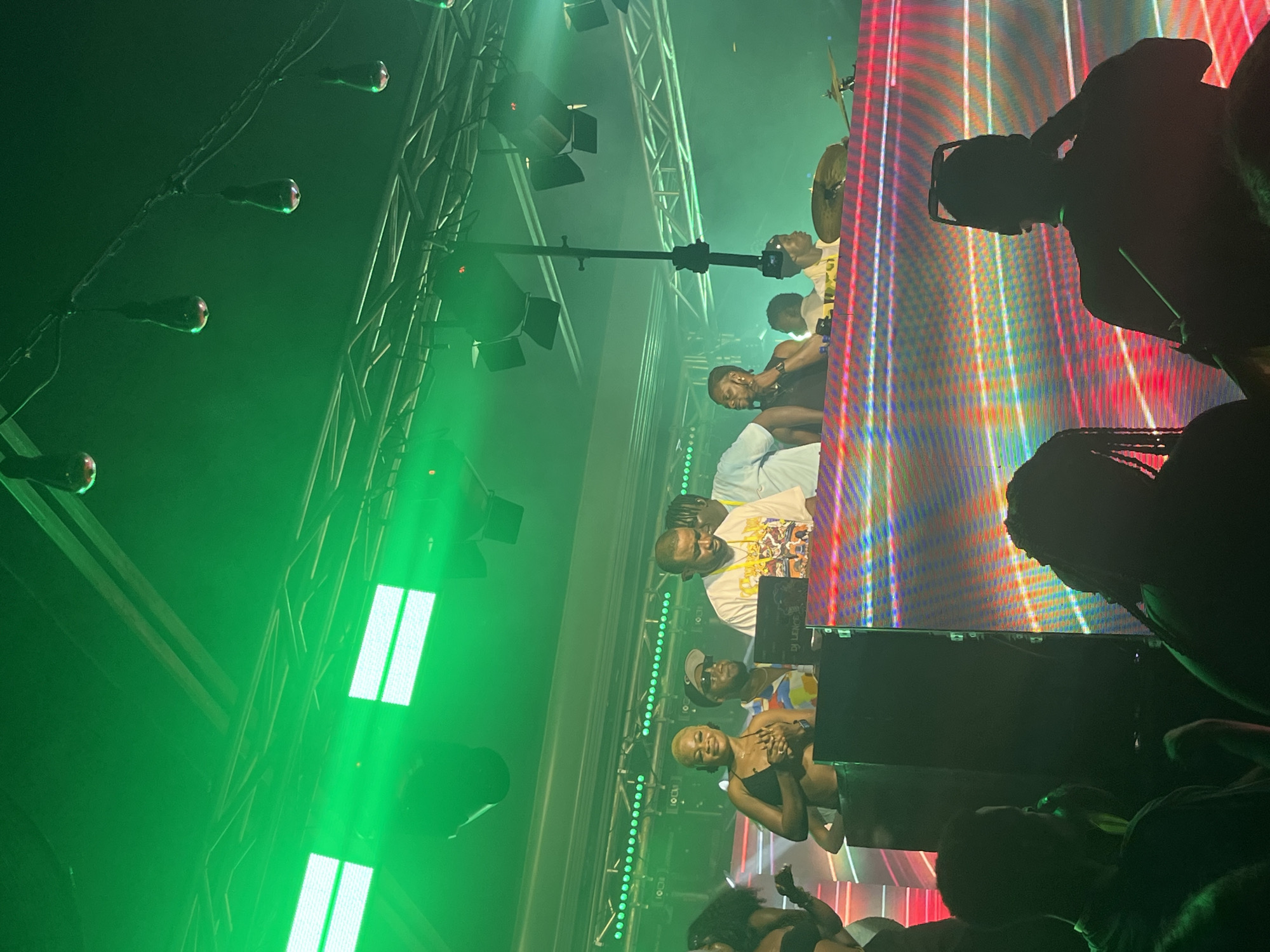 The image depicts a lively event at a venue, possibly a club or concert hall, in Howayi, Nigeria. The scene is dominated by vibrant green lighting and stage lights. In the background, a DJ setup is visible, with a DJ at the console. Several individuals are on stage, some appearing to be performers or guests. A woman with a blonde hairstyle is prominently featured in the foreground of the stage area, smiling and engaging with the event. Behind her, a man wearing a white t-shirt with a graphic print and a cap is standing. Another man, wearing sunglasses, is next to him. A drummer is also present on the stage, with cymbals visible. In front of the stage, the audience is visible, with some individuals silhouetted against the colorful LED screen displaying abstract patterns. The overall atmosphere suggests a dynamic and energetic gathering.