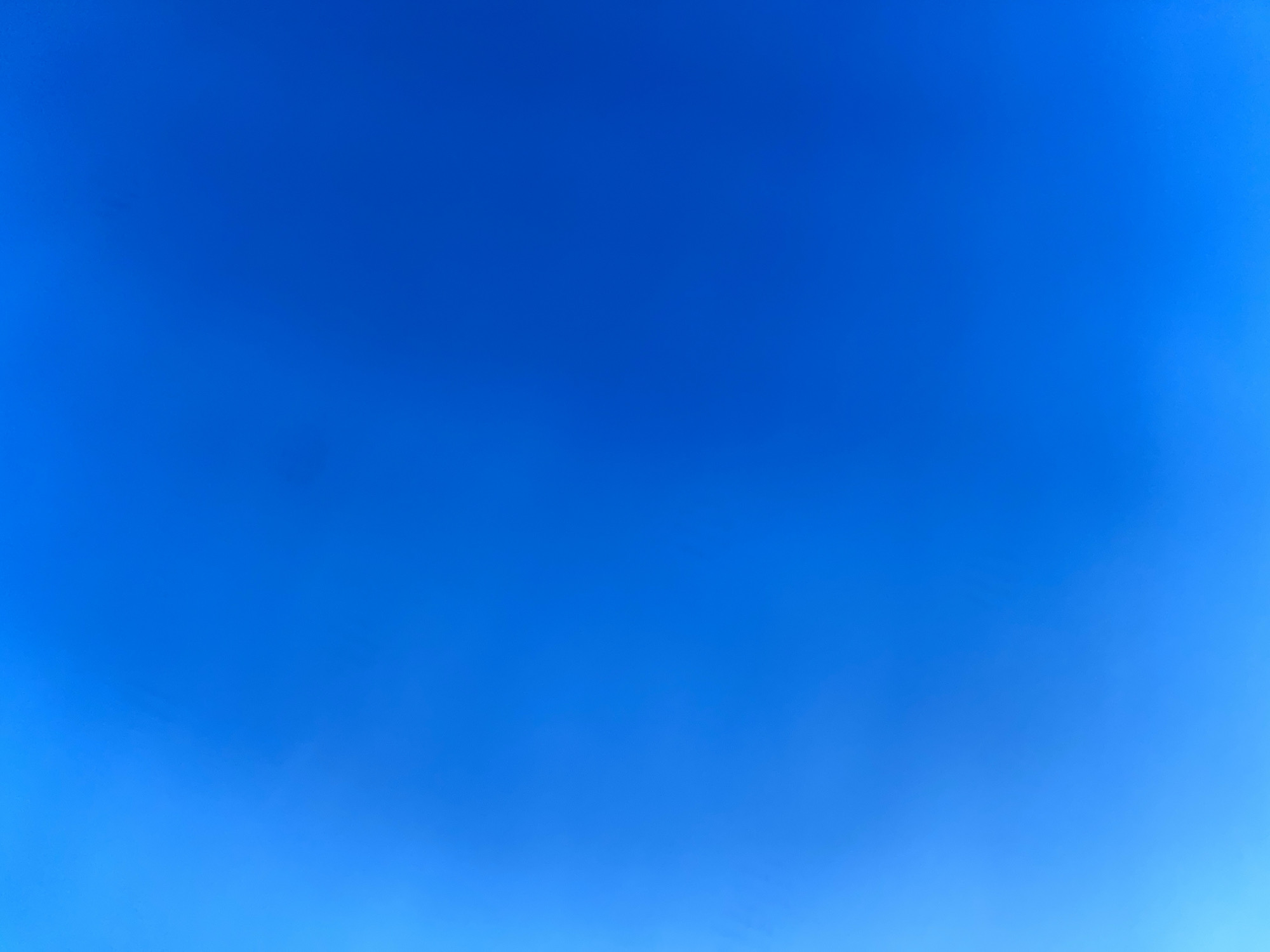 This is a full-frame, high-angle, outdoor shot of a clear blue sky. There is a subtle gradient of color, with the sky appearing lighter on the left side of the frame and a deeper blue towards the right and top. No objects or people are visible in the frame. The image depicts the sky over the village of Kaliuzhyntsi, Ukraine.