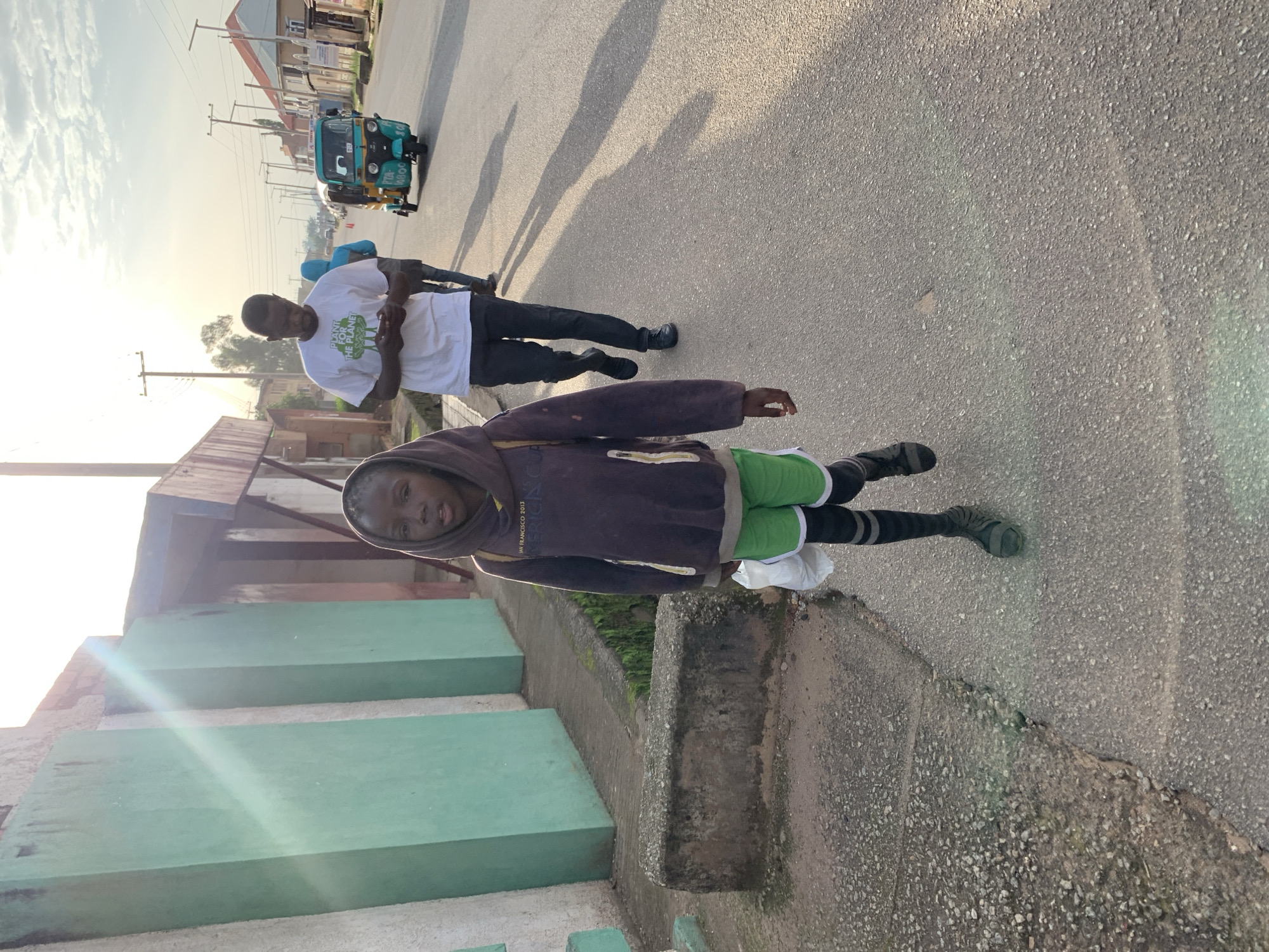 In Jos, Nigeria, a young child with dark skin and wearing a dark hooded sweatshirt over green shorts, black and grey striped socks, and dark sandals walks towards the camera on a paved road. The child is carrying a white plastic bag in their right hand. Behind the child, a man in a white t-shirt with a green logo reading "PLANT FOR THE PLANET" and dark pants walks away from the camera, looking at his phone. Another person in a blue hooded jacket is visible further back. A teal and yellow auto-rickshaw is driving on the road in the background, followed by other vehicles. The sun is low in the sky, casting long shadows and creating lens flare on the left side of the image, illuminating the scene. Buildings line the street, with a green-painted wall on the left and other structures visible on the right.