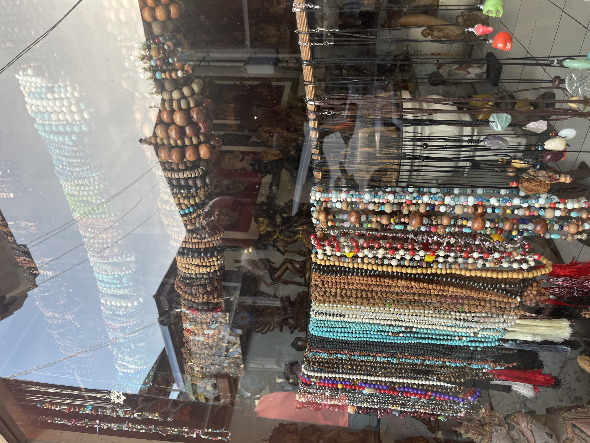A display window in Ubud District, Indonesia, exhibits an array of jewelry. Beaded bracelets and necklaces of varying colors and materials are stacked densely. Some necklaces include tassels. Carved wooden objects are visible behind the jewelry. A glass surface reflects the street.