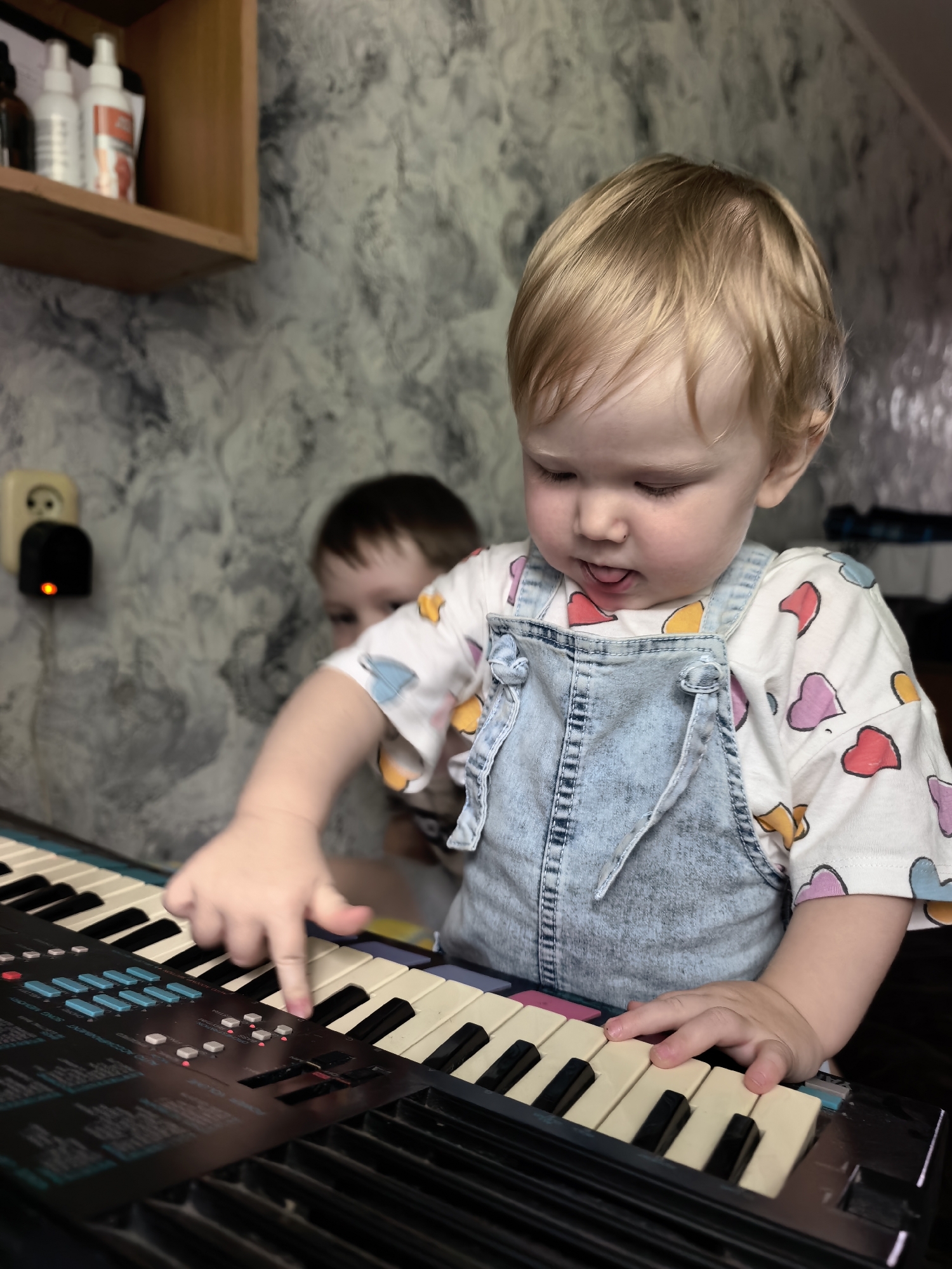 A young child with blond hair, wearing a light blue denim overall and a white t-shirt adorned with colorful heart patterns, is playing a black electronic keyboard. The child's tongue is slightly sticking out as they focus on pressing the keys with their right index finger. In the background, another child is partially visible, blurred and looking towards the keyboard. To the left of the main subject, a beige shelf holds several bottles, and a wall socket with a black adapter is mounted on a textured grey wall. The overall scene suggests a domestic indoor environment.