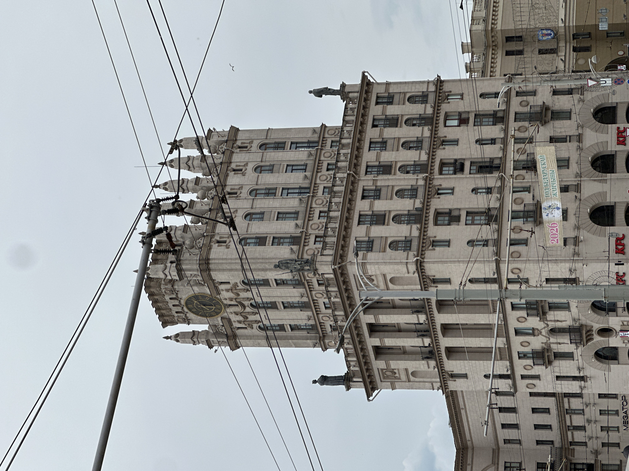 The image captures a tall, light-colored building in the Stalinist Empire style, identified as one of the "Gates of Minsk" towers, located at Pryvakzalnaja Square in front of the Minsk Passenger railway station in Minsk, Belarus. The multi-story structure features a prominent clock tower with a large circular clock face displaying Roman numerals. Two statues of figures, one appearing to be a worker and the other possibly a farmer or soldier, stand on the upper terrace of the clock tower. Another statue, depicting a cloaked figure, is positioned on a lower terrace on the left side of the building.
The building exhibits numerous arched and rectangular windows, many with small, ornate metal balconies. Decorative architectural elements such as cornices, balustrades, and relief work adorn its facades. The ground level shows commercial establishments, including a partially visible "KFC" sign on the right and "MEGATOP" on the left.
The foreground is dominated by multiple grey and black overhead utility wires, characteristic of trolleybus or tram lines, which run horizontally and diagonally across the frame, partially obscuring the building and the sky. A grey utility pole is prominently visible in the lower right and middle. A banner, partially visible, hangs horizontally in the lower mid-section with the Cyrillic text "ГОД БЕЛАРУСКАЙ ЖАНЧЫНЫ" (Year of Belarusian Women) and the number "2026".
The sky is overcast and light grey. A single bird, possibly a gull, is visible in flight in the upper right quadrant of the image. A portion of an adjacent building, also of similar architectural style, is visible to the right, beyond the utility pole.