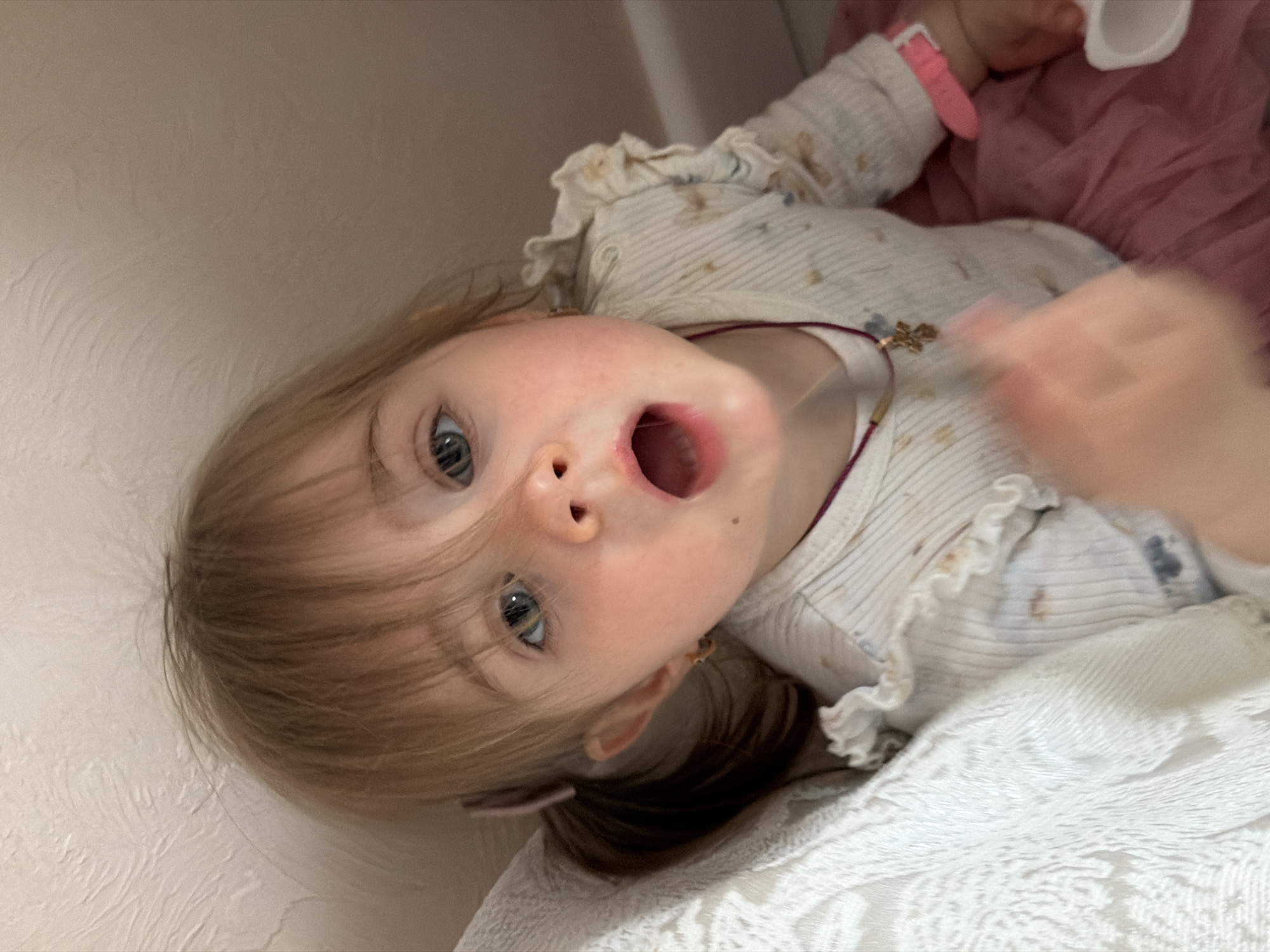 This image presents a close-up, angled portrait of a young child, likely a toddler, in an indoor setting. The child, with light blonde or light brown hair that falls across their forehead, has wide, striking blue eyes that gaze directly at the viewer. Their mouth is open wide, suggesting an expression of surprise, a yawn, or the act of making a sound.
The child is dressed in a light-colored, possibly cream or white, long-sleeved top featuring ruffled details on the shoulders and a fine striped texture with subtle markings or patterns. A small, golden-colored charm necklace is visible around their neck, and a pink watch adorns their left wrist. A reddish-pink fabric, possibly a dress or skirt, is visible on their lower body. The child's right hand (from their perspective) is in motion and appears blurry, adding a dynamic element to the image.
The background consists of a plain, light-colored, textured wall against which the child is reclining or lying. In the foreground, a white, textured blanket or fabric is visible. The photograph is taken from a slightly elevated and diagonal perspective, making the child's face the central and dominant feature. There is no visible text in the image. The scene is set in Rossiyskiy, Russia, conveying a domestic atmosphere.
