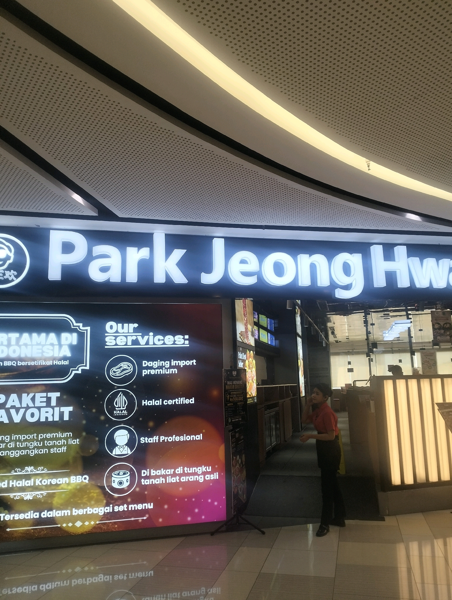 The image shows the exterior of a "Park Jeong Hwa" restaurant, which appears to be a Korean BBQ establishment. The restaurant's name is prominently displayed on a large, illuminated sign. Below the sign is a large, digital advertisement board that showcases various services offered by the restaurant, such as "Daging import premium," "Halal certified," "Staff Profesional," and "Di bakar di tungku tanah liat arang asli." The advertisement also highlights that this is the "Pertama di Indonesia" (First in Indonesia) for Halal Korean BBQ, and that set menus are available.
A young female employee, wearing a red shirt and black pants, is standing near the advertisement, looking towards the left side of the frame and appears to be adjusting her hair or earpiece. She is standing on a tiled floor. The environment suggests an indoor setting, possibly a mall or a commercial complex, given the modern ceiling design with integrated lighting and perforated panels. The lighting is bright and artificial, illuminating the restaurant's facade and the advertisement. The reflection of the ceiling and lights is visible on the floor. The overall scene is well-lit and presents a clean, professional image of the restaurant. It is implied this is in Surabaya, Indonesia due to the Indonesian text and "Halal Indonesia" certification.