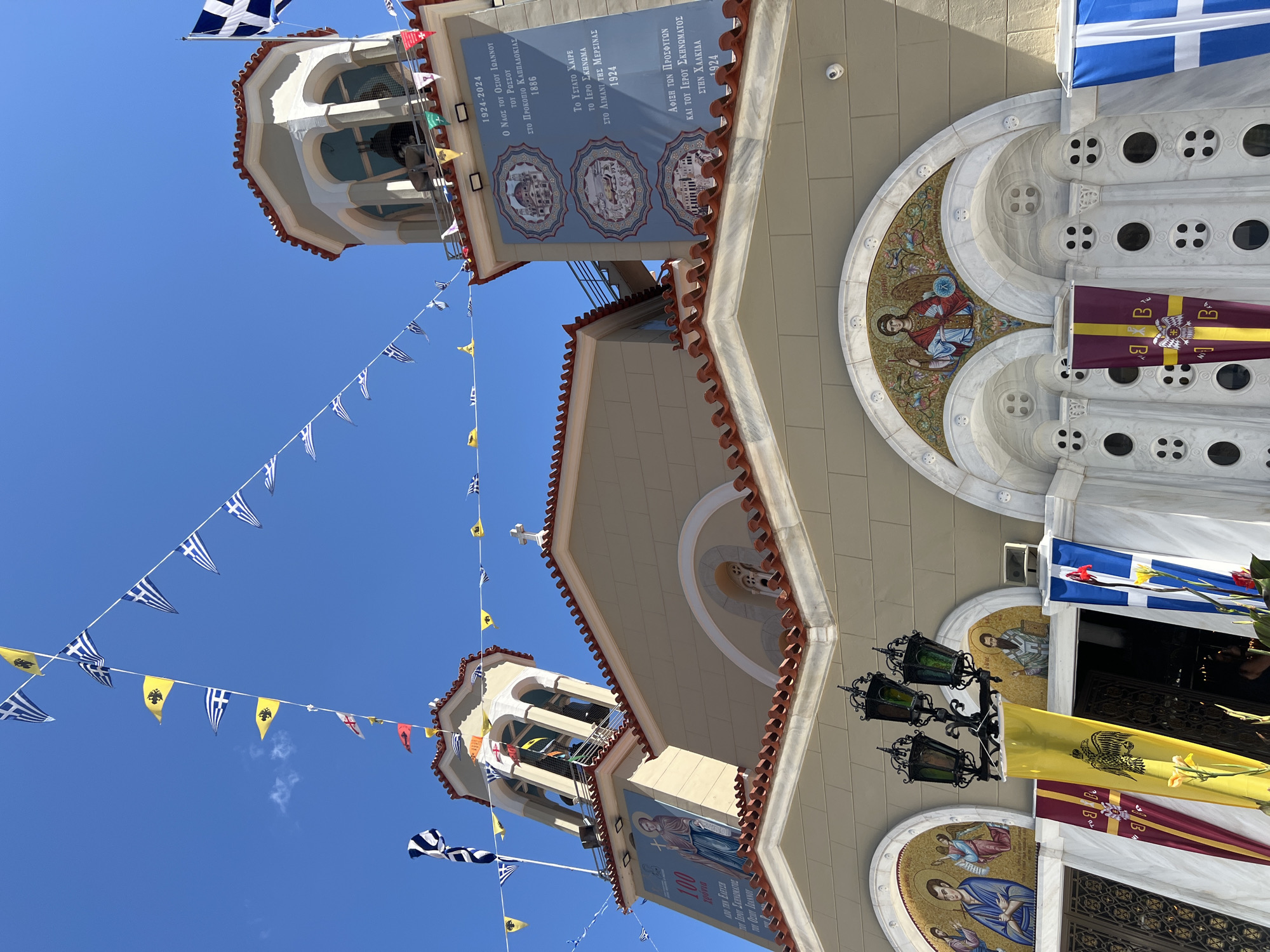 The image shows the exterior of a church in the city of Prokopi, Greece, under a clear blue sky. The church has a light beige facade with a red-tiled roof. Two bell towers are visible, adorned with flags and pennants. One tower has a mural of a saint and text that reads "1924-2024" and "Ο ΝΑΟΣ ΤΟΥ ΟΣΙΟΥ ΙΩΑΝΝΟΥ ΤΟΥ ΡΩΣΟΥ ΣΤΟ ΠΡΟΚΟΠΙΟ ΚΑΠΠΑΔΟΚΙΑΣ 1886". String lights with small Greek flags and other pennants are strung across the sky and between the towers.
In the foreground, the entrance of the church is decorated with large banners and religious mosaics above the arched doorways. One mosaic depicts an angel. The church features multiple arched windows and decorative architectural elements. A lamppost with three lanterns stands in front of the entrance. Several people are partially visible entering or exiting the church. The overall atmosphere suggests a religious celebration or anniversary.