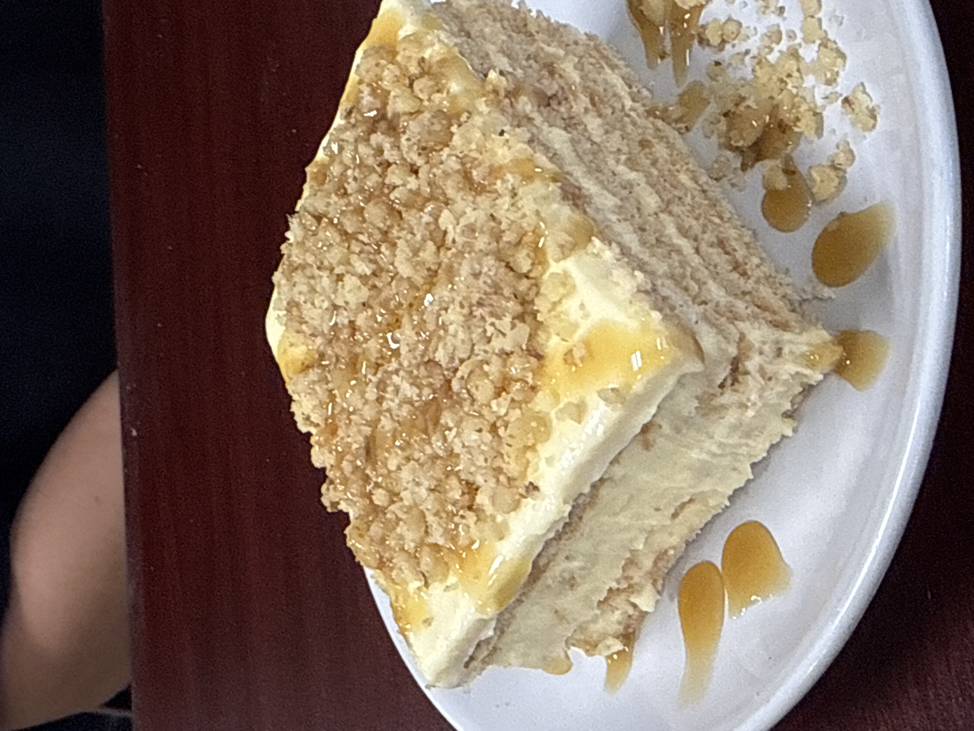 A slice of layered cake is presented on a white plate. The cake appears to have a light-colored filling between the layers, and the top is covered with a crumbly topping, possibly nuts, drizzled with a golden syrup. The plate also has a few droplets of the same syrup. The scene is set on a dark red surface with a portion of a black garment and arm visible on the left edge of the frame. The location cannot be determined to be in Des Plaines, United States of America based on visual information alone.
