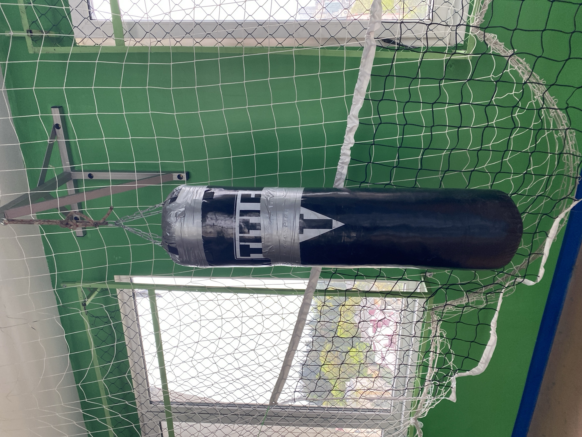 The image depicts a black "TITLE" branded punching bag hanging from a silver metal frame via chains, against a backdrop of green wall partially covered by a net. Light from a window illuminates the scene. It is not possible to confirm the location as Sochi, Russia based solely on the visual information provided.