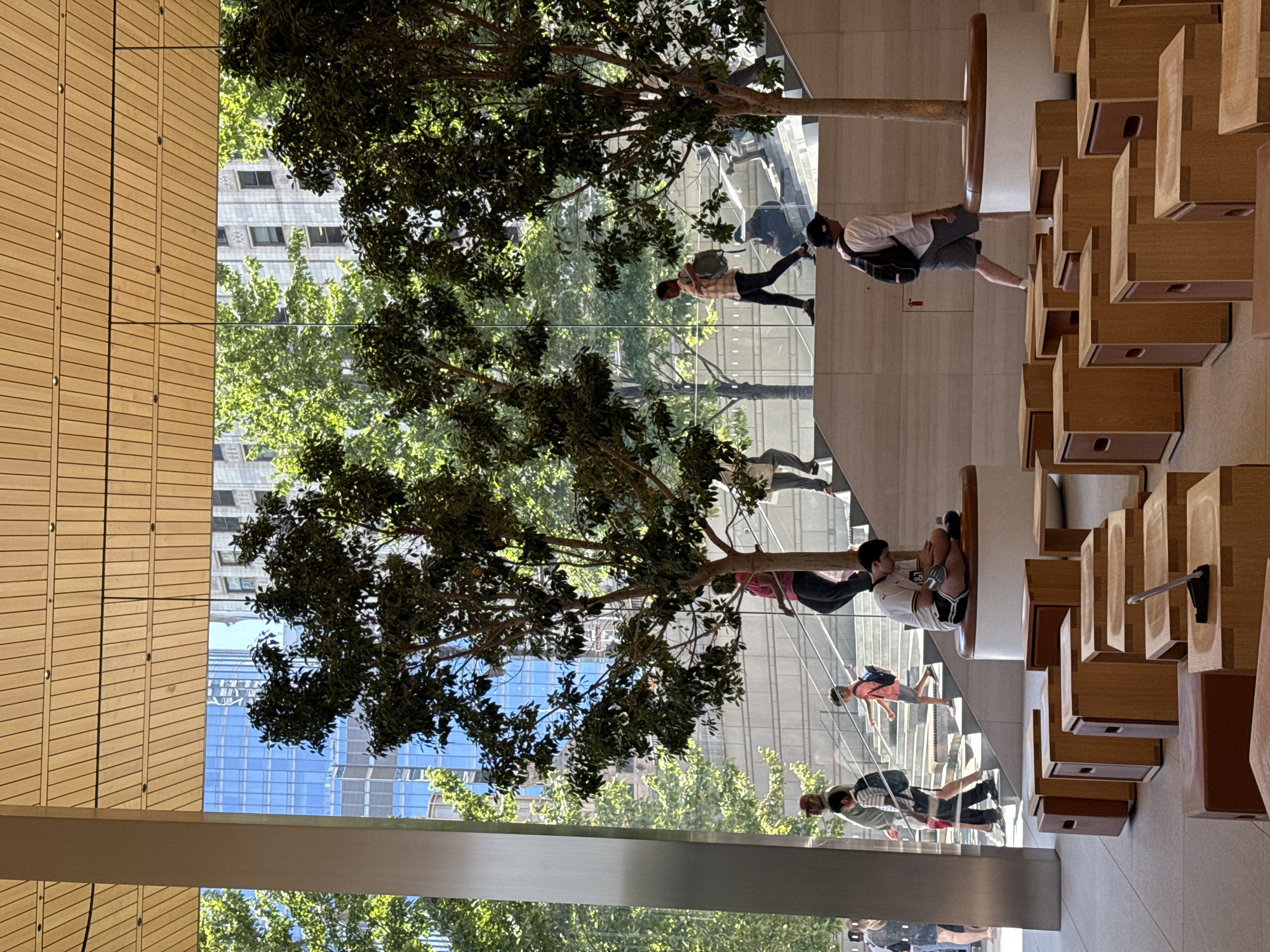 This image depicts the interior of an Apple Store in Chicago, Illinois, USA, characterized by its modern architectural design. The scene features a multi-level layout with glass walls offering views of trees and city buildings. In the foreground, numerous wooden cubic seating units are arranged on a tiled floor. Several people are present, walking on stairs or sitting on benches. One person is seated on a circular bench in the middle ground, looking down. Another individual wearing a backpack is walking towards the right. Further back, people are ascending a wide staircase. The overall environment suggests a bright, open, and contemporary retail space.