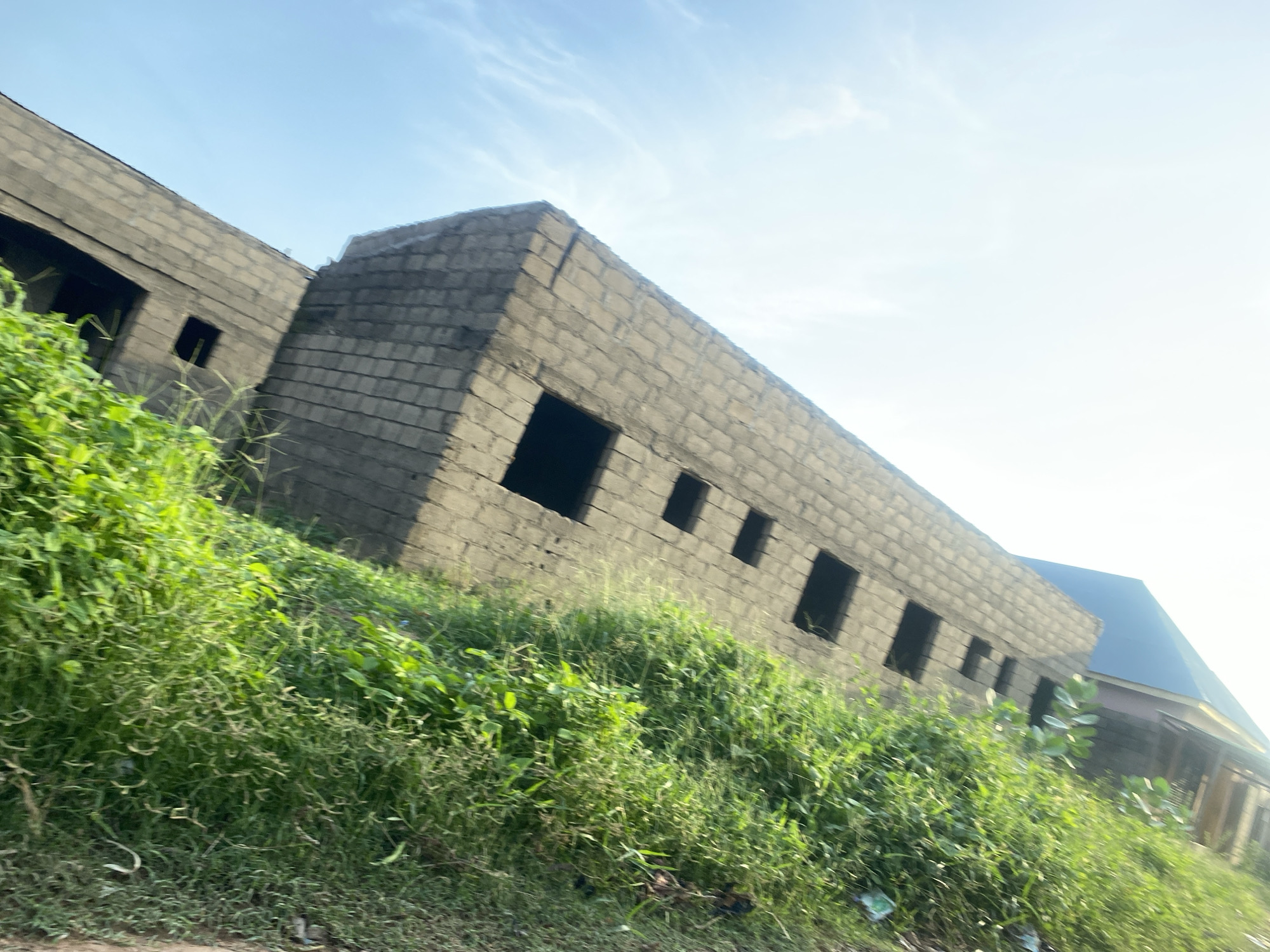 The image depicts an outdoor scene in Jalingo, Nigeria, under a clear, bright sky suggesting daytime with fair weather. The primary subjects are two unfinished buildings constructed from gray cinder blocks. The building in the foreground is long and low, with several dark, rectangular openings that appear to be window or doorway frames. This structure is partially obscured by an abundance of tall, green grass and foliage that grows densely on a sloped embankment in front of it.
Behind the first building, a second, slightly taller building is visible, also constructed of cinder blocks and appearing to be in a similar state of incompletion. This second building has a dark, sloped roof, possibly made of metal. The scene is devoid of people or any discernible activity. The environment appears somewhat overgrown, with the vegetation encroaching on the construction site. The overall impression is one of ongoing construction or possibly abandoned building projects. There is no visible text in the image.