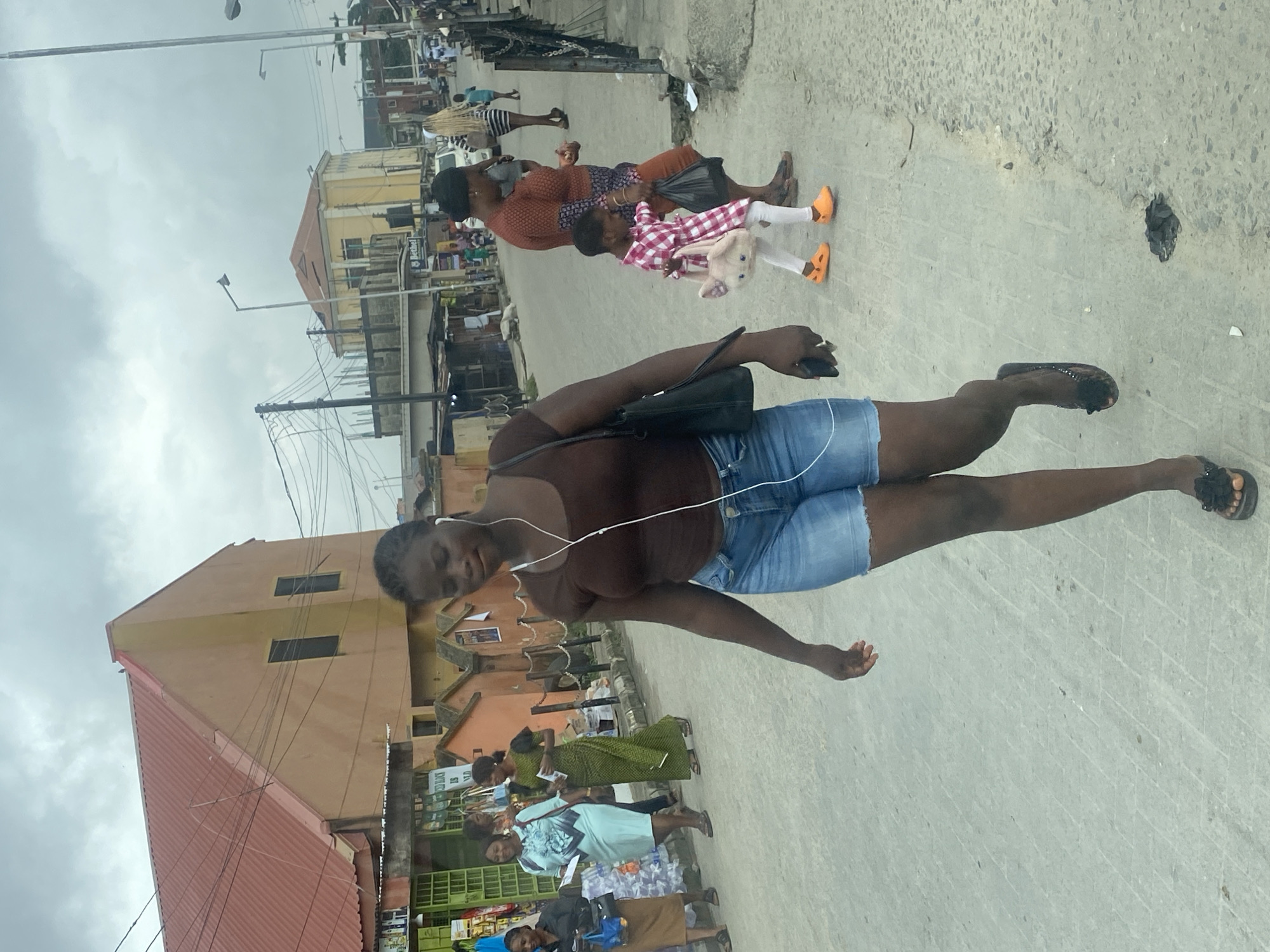 The image depicts a street scene in an urban environment. In the foreground, a young woman with dark skin is walking towards the viewer, wearing a brown sleeveless top and denim shorts. She has white earbuds in her ears and is carrying a black shoulder bag. Her feet are bare, wearing black flip-flops with flower embellishments.
Several other individuals are visible in the background. To the left, a woman in a light blue dress is walking away from the viewer, holding papers. Beside her, a woman in a green patterned dress is also walking away. Further back, a woman in a rust-colored patterned top and orange pants is walking, holding a black plastic bag. A young child, dressed in a pink and white checkered dress and white leggings, is walking beside the woman in the rust-colored top, holding a pink bag shaped like a cat's head and wearing bright orange shoes. In the distance, a woman with blonde hair in a striped dress is walking.
The street is paved with concrete. Buildings line the street, with one prominently featuring a red-tiled roof and an orange-yellow facade. Other buildings are visible further down the street, some appearing to be shops with green shutters. Power lines and streetlights are present in the scene. The sky is overcast. The overall impression is a busy street in what appears to be a commercial or residential area of a city.
